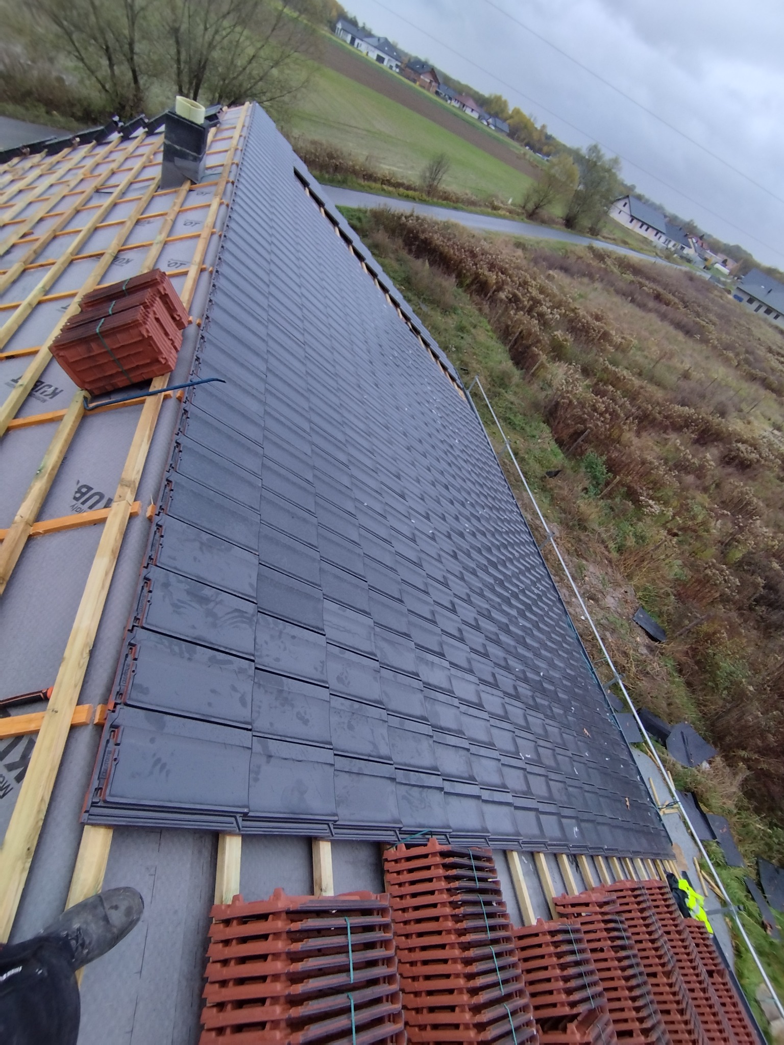 Układanie grafitowej dachówki na konstrukcji drewnianej dachu, widoczne stosy czerwonej dachówki w tle krajobraz z polami i zabudowaniami.