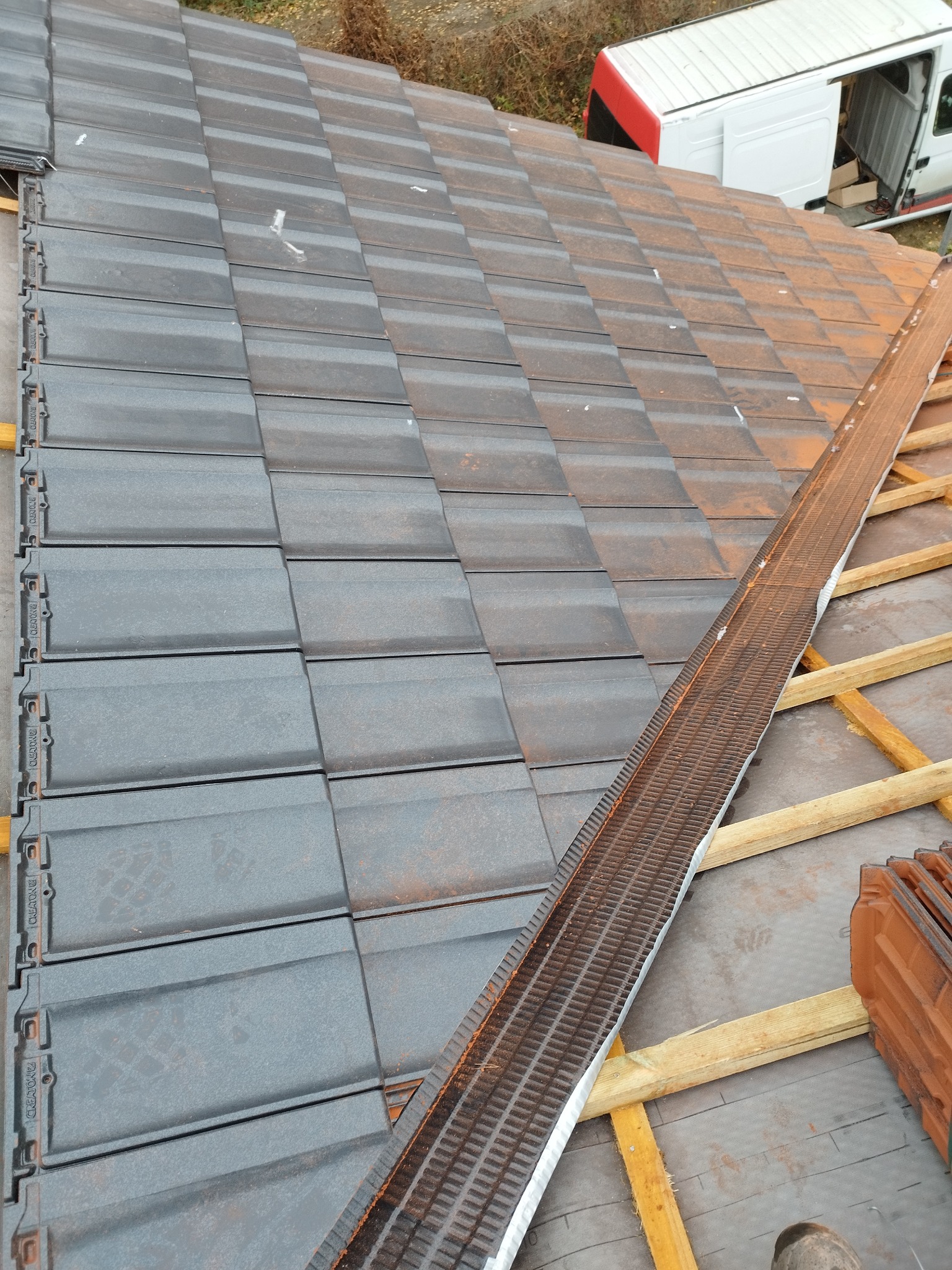 Fragment dachu z częściowo ułożoną, szarą dachówką ceramiczną na drewnianej konstrukcji, widoczna membrana dachowa i elementy wentylacyjne, w tle biały samochód dostawczy.