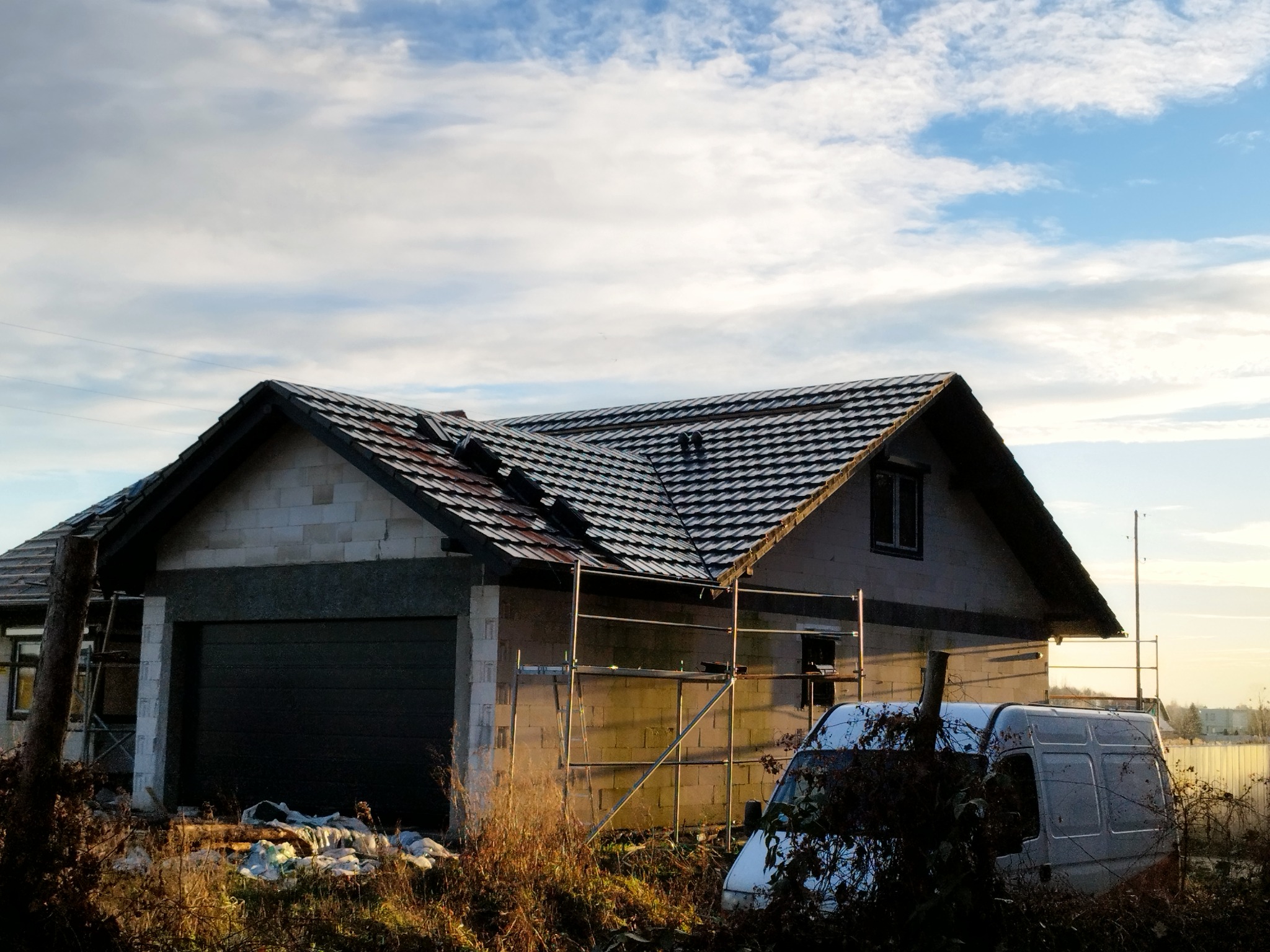 Dom jednorodzinny w trakcie budowy z ciemną dachówką, widoczne rusztowania i zaparkowany van, nieukończona elewacja z bloczków.