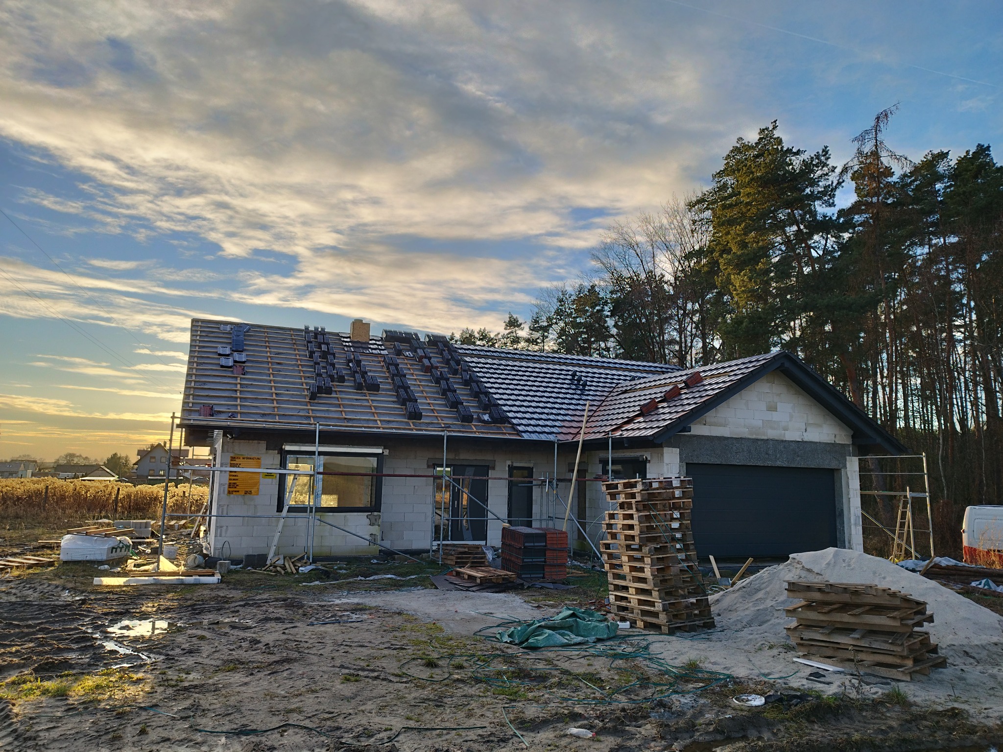 Niedokończony dom jednorodzinny z częściowo ułożonym ciemnoszarym pokryciem dachowym, drewnianą konstrukcją dachu i stertami dachówek na połaci, otoczony błotnistym terenem budowy i drzewami w tle.