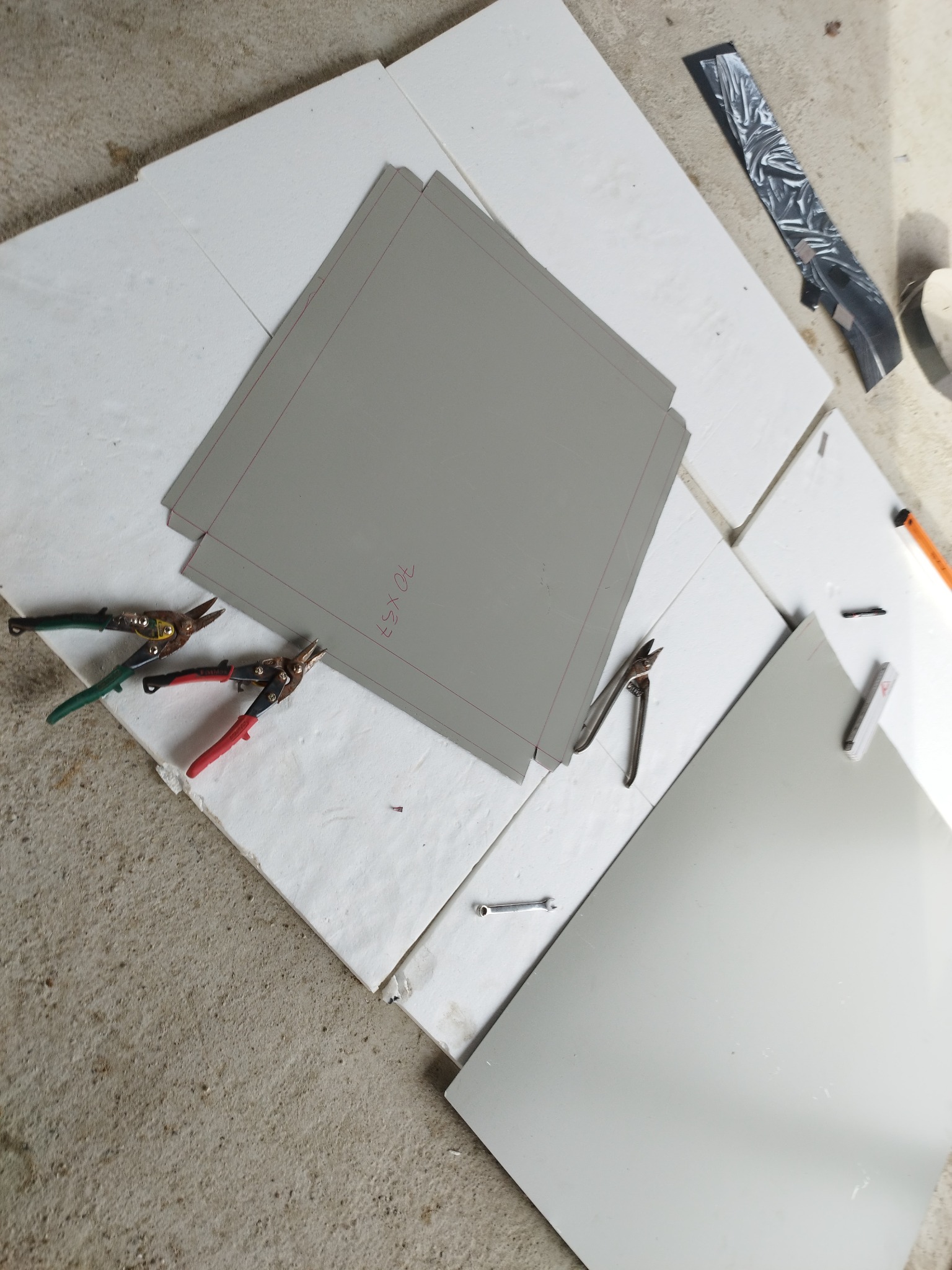 Przygotowanie arkuszy blachy z naniesionymi wymiarami 70x53 cm, nożyce do blachy z zielonymi i czerwonymi uchwytami oraz inne narzędzia na białych płytach styropianowych, podłoże betonowe.