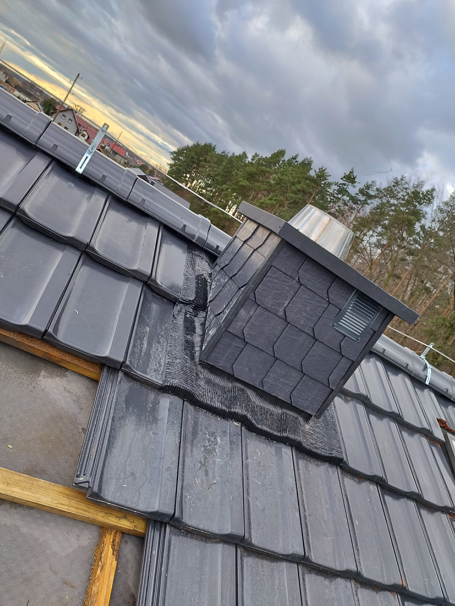 Montaż komina wentylacyjnego z łupka na dachu pokrytym grafitową dachówką ceramiczną, widoczne fragmenty drewnianej konstrukcji więźby dachowej i metalowe uchwyty mocujące.