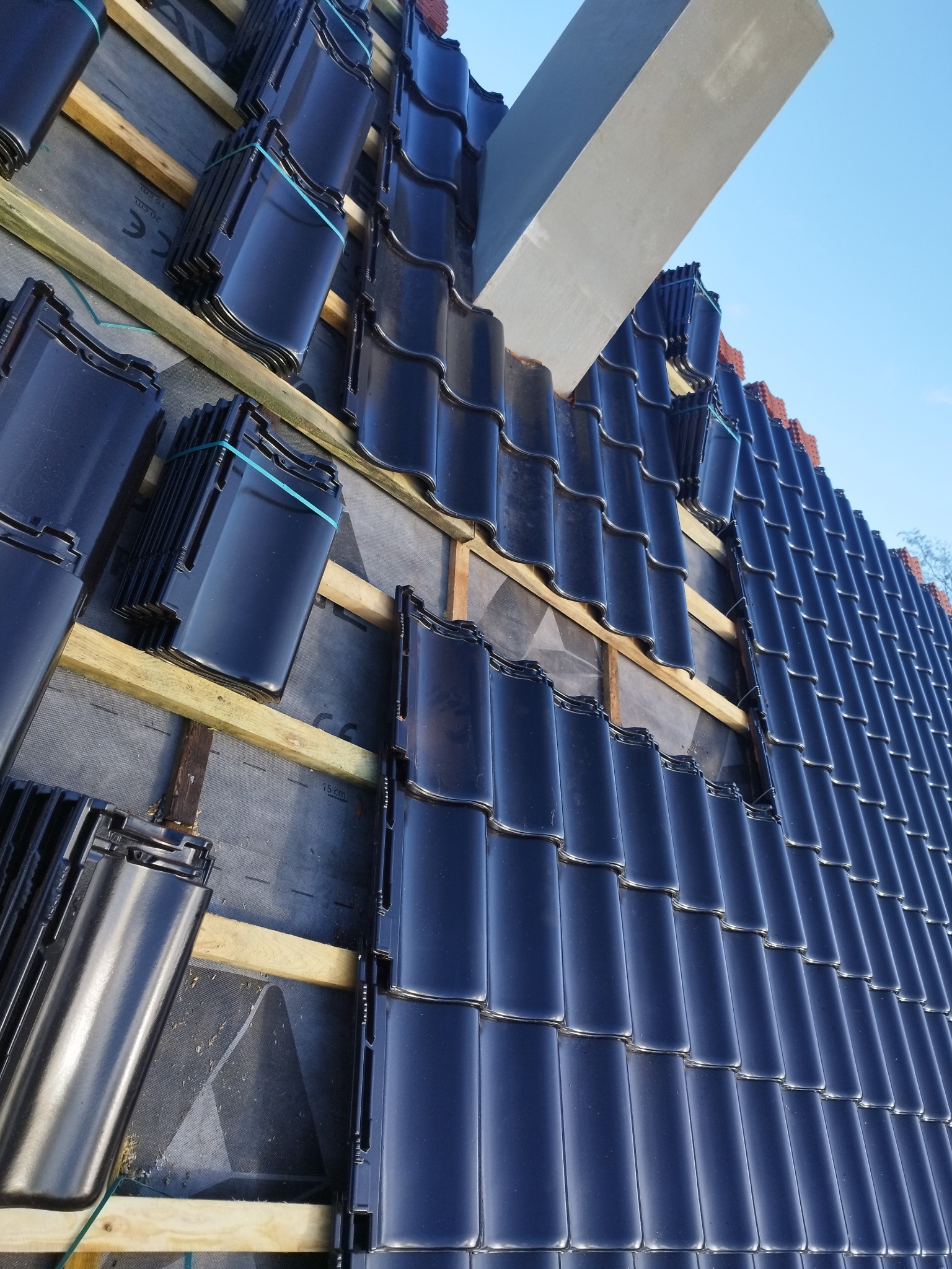 Układanie błyszczącej, czarnej dachówki karpiówki na membranie dachowej, widoczna drewniana konstrukcja łaty i fragment komina, dach w trakcie prac.