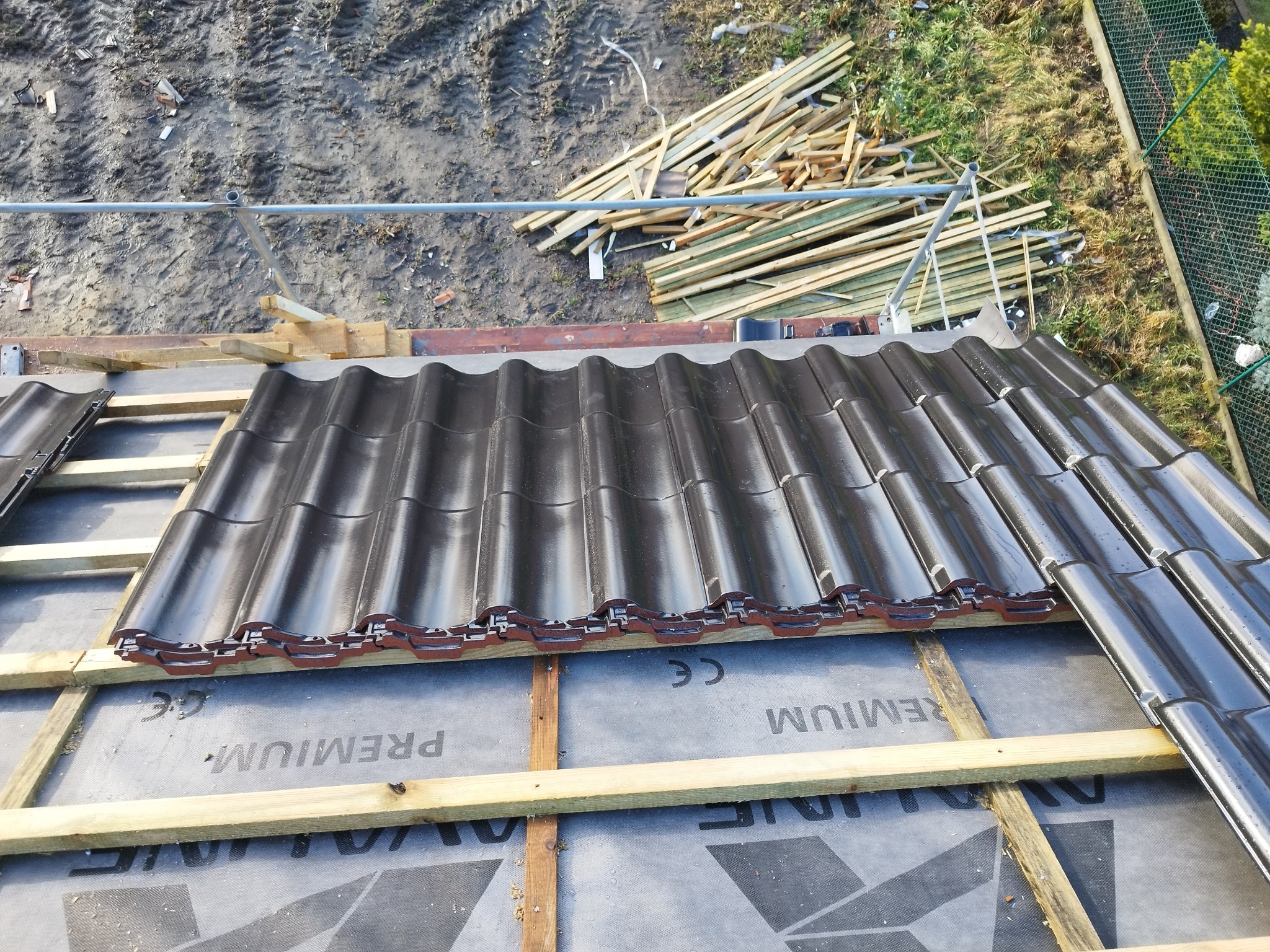 Układanie ciemnej dachówki ceramicznej na łatach i membranie dachowej, widoczny fragment rusztowania i stos desek w tle.