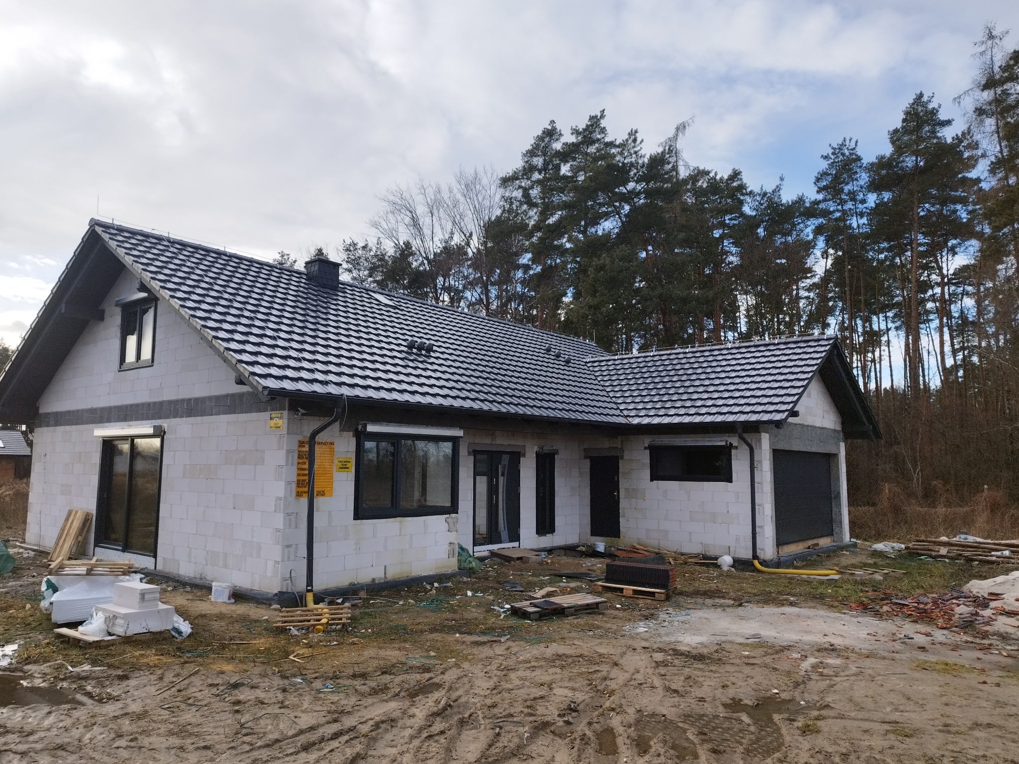 Nowy dom jednorodzinny w trakcie budowy, z ciemną dachówką karpiówką na dachu i jasnymi ścianami z bloczków, widoczne materiały budowlane i rynna