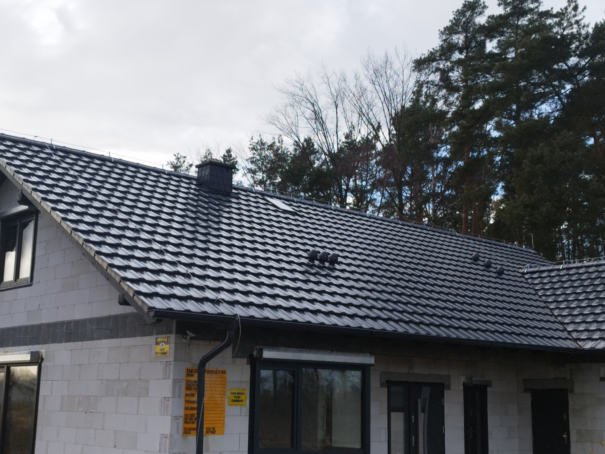 Dom w stanie surowym zamkniętym z nowym, ciemnym dachem z dachówki ceramicznej, widoczne kominki wentylacyjne i rynna, na tle drzew.