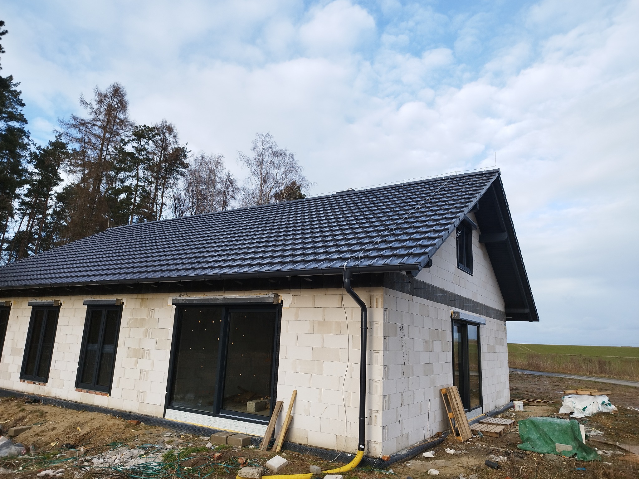 Dom w trakcie budowy z czarnym dachem z blachodachówki, widoczne okna z czarnymi ramami, ściany z białego bloczka, orynnowanie w kolorze czarnym, w tle drzewa i pole.