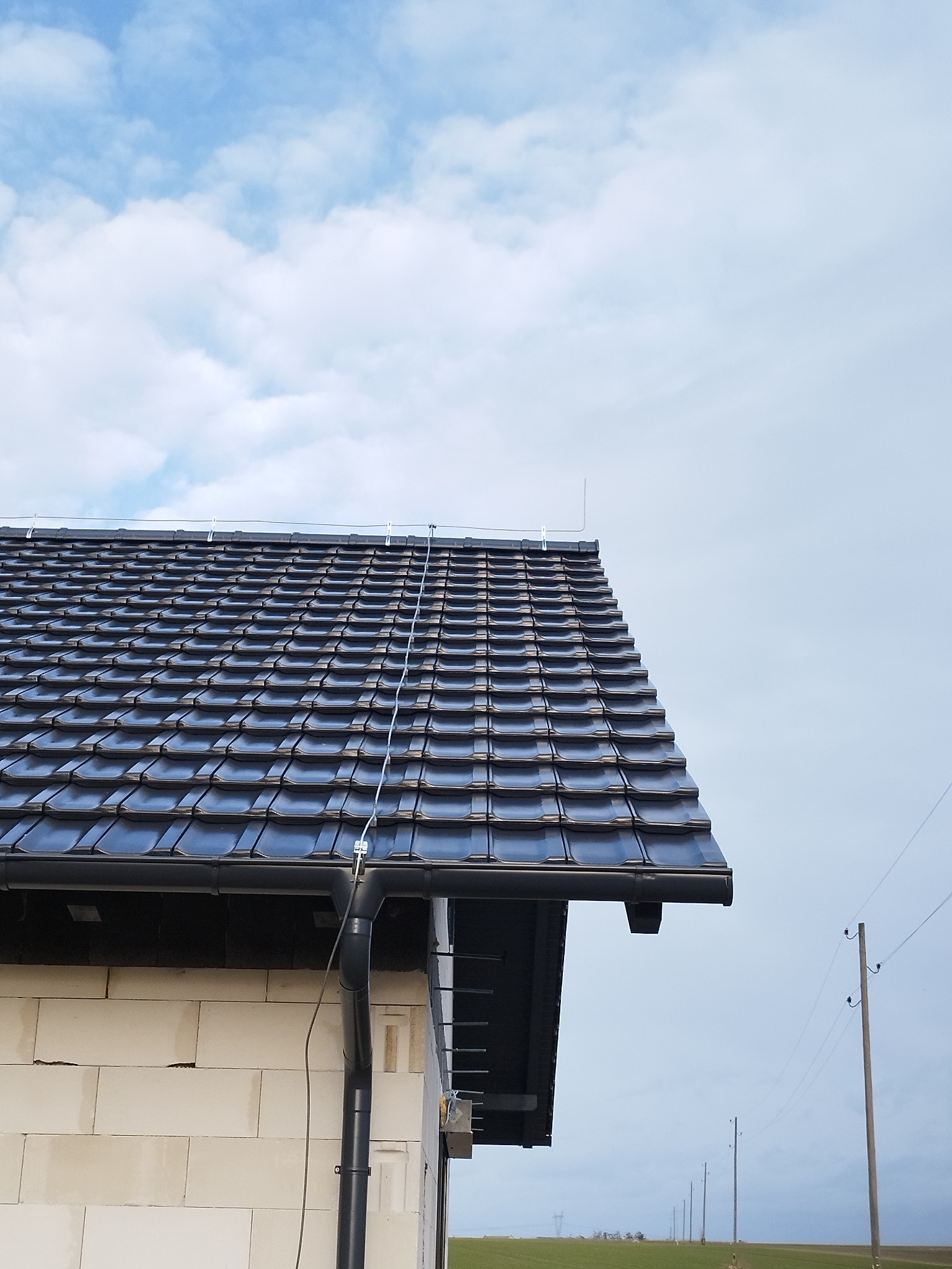 Nowy dach z czarnej dachówki ceramicznej na budynku w trakcie budowy, widoczne rynny i przewody odgromowe, tło stanowi błękitne niebo z chmurami.