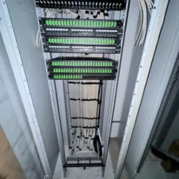 Szafa serwerowa z uporządkowanymi kablami sieciowymi, patch panele z zielonymi portami, widok z dołu w trakcie instalacji.