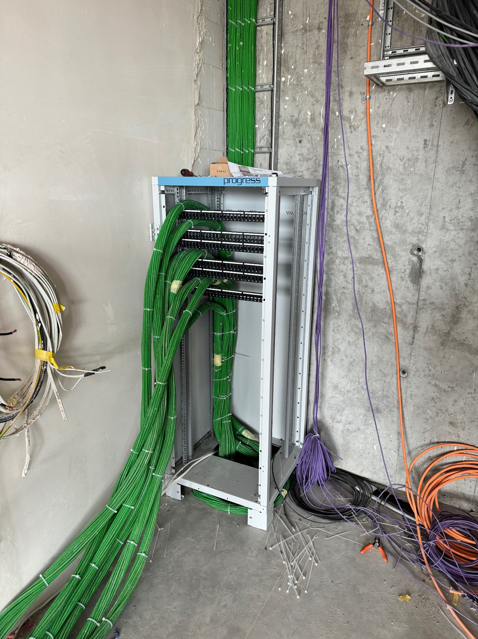 Szafa serwerowa z podłączonymi kablami w kolorach zielonym, fioletowym i pomarańczowym, częściowo uporządkowanymi, z widocznymi narzędziami do cięcia kabli i opaskami zaciskowymi na surowym...