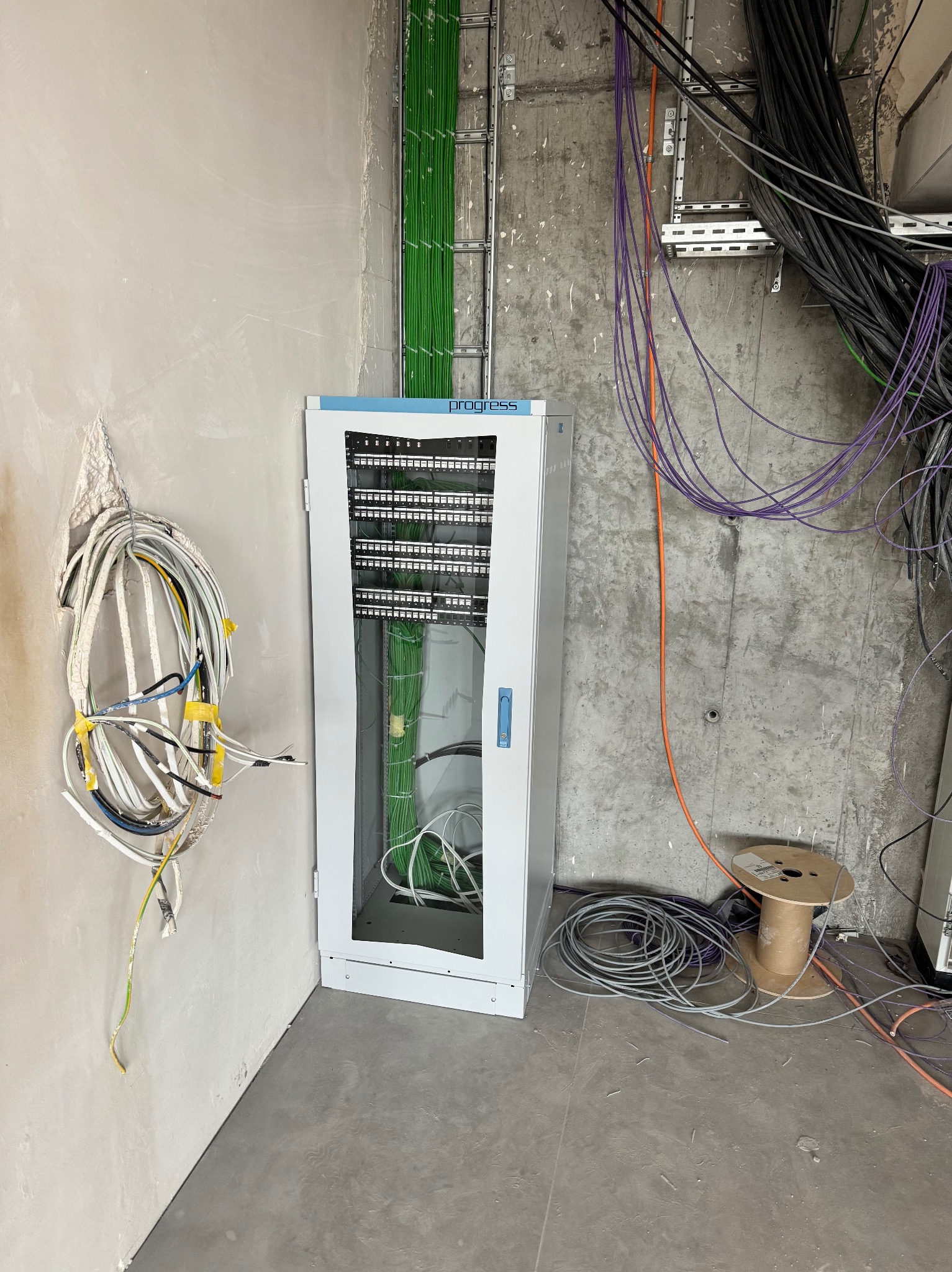 Szafa rackowa z patch panelem i okablowaniem w surowym pomieszczeniu, przewody ułożone w korytkach, zwój kabli przymocowany do ściany, bęben kablowy na podłodze.