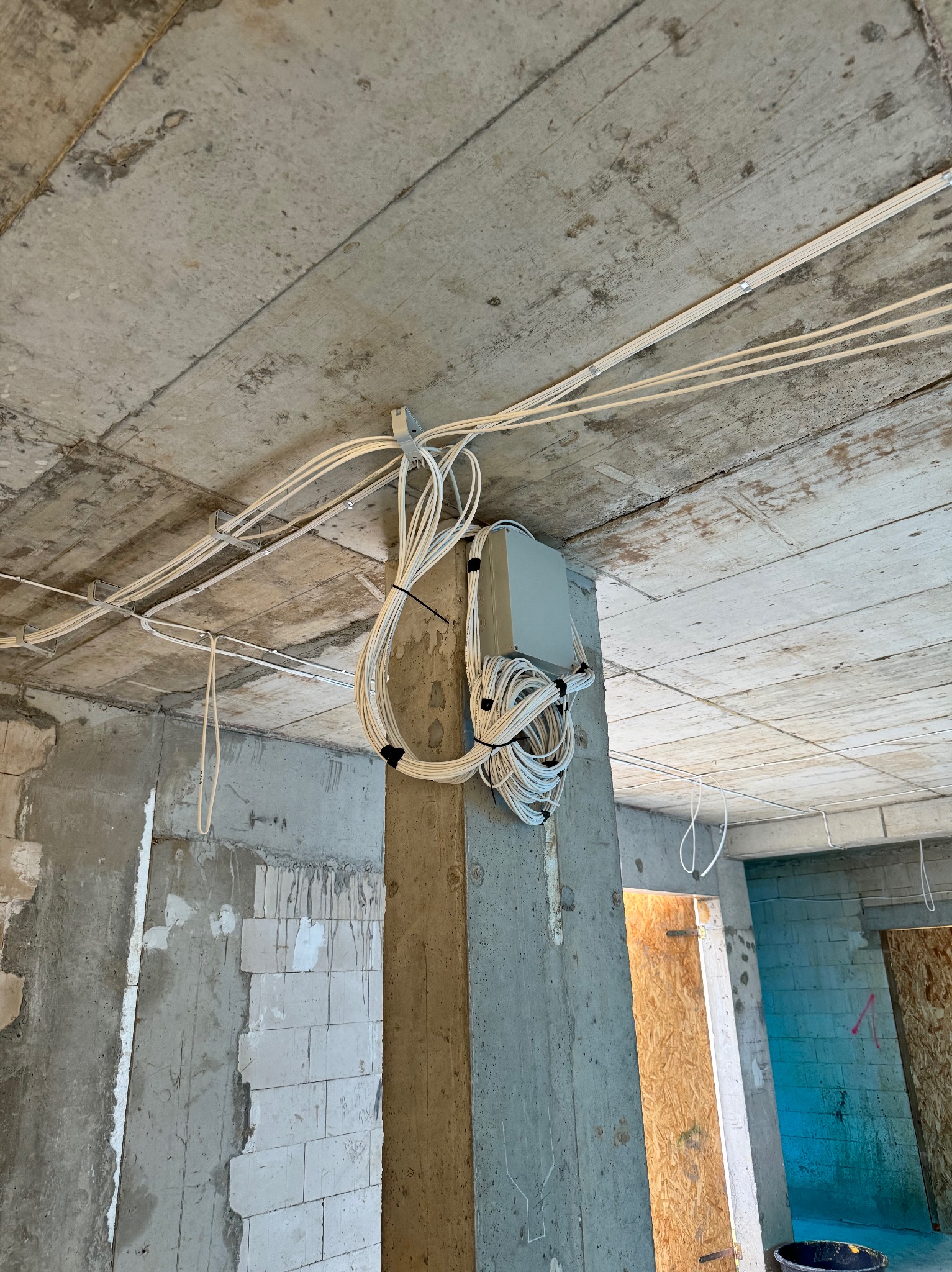 Surowe wnętrze w trakcie remontu z widocznymi przewodami elektrycznymi mocowanymi do betonowego sufitu i kolumny, szara skrzynka elektryczna przytwierdzona do filaru, widoczne kable ułożone w pętle...