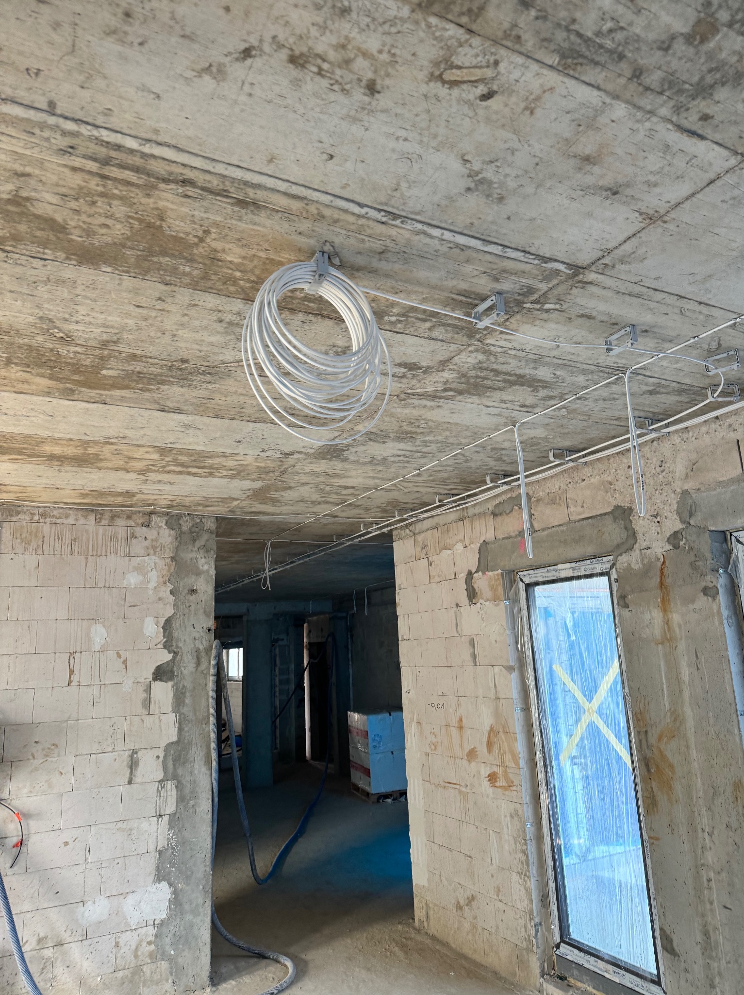 Wnętrze w stanie surowym z widoczną instalacją elektryczną w trakcie montażu: zwój kabli podwieszony do sufitu, przewody prowadzone po betonowym stropie, ściany z białego pustaka.