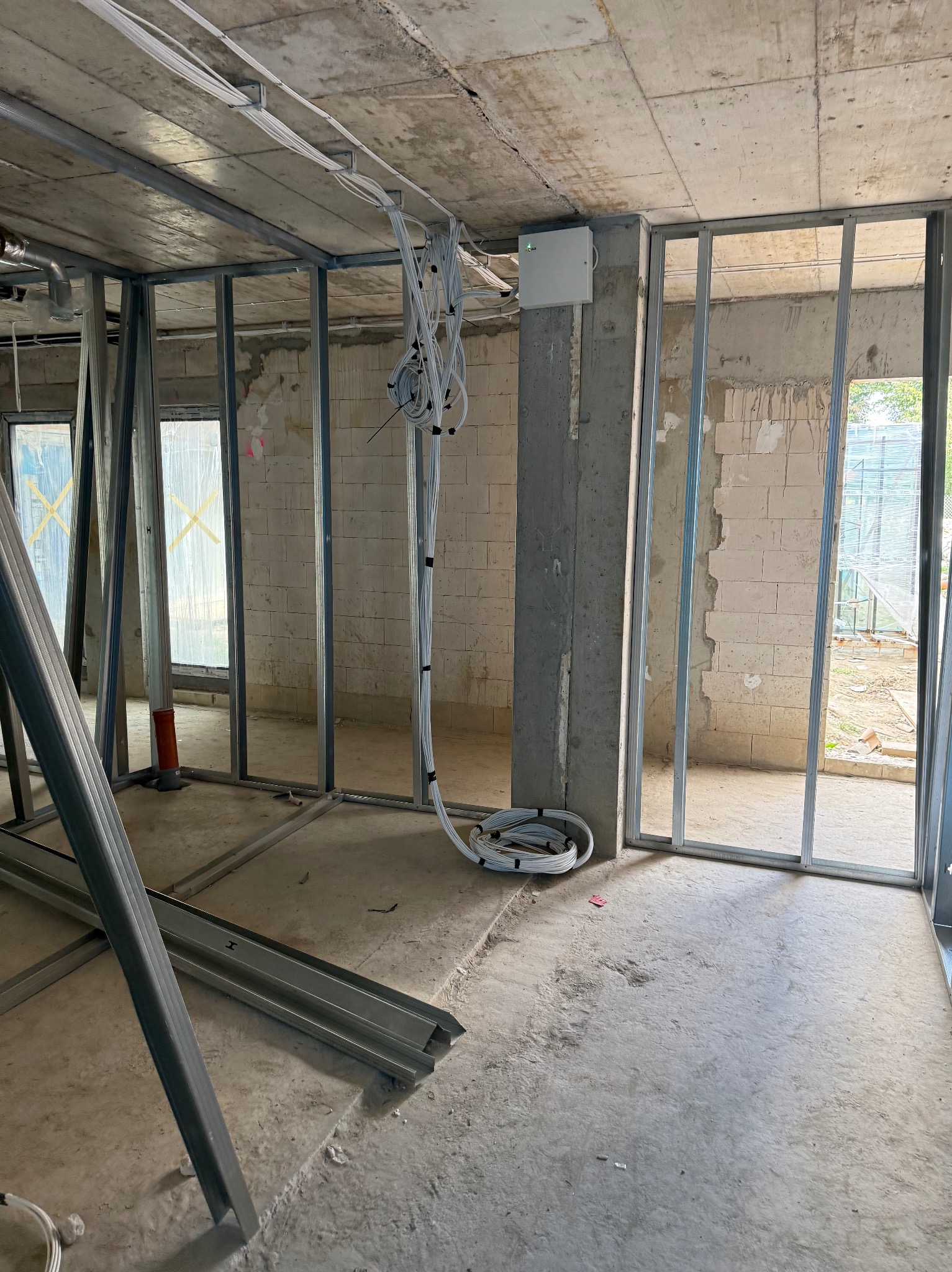 Wnętrze w stanie surowym z widocznymi metalowymi profilami ścian działowych, wiązkami kabli elektrycznych pod sufitem i puszką elektryczną na betonowym słupie.