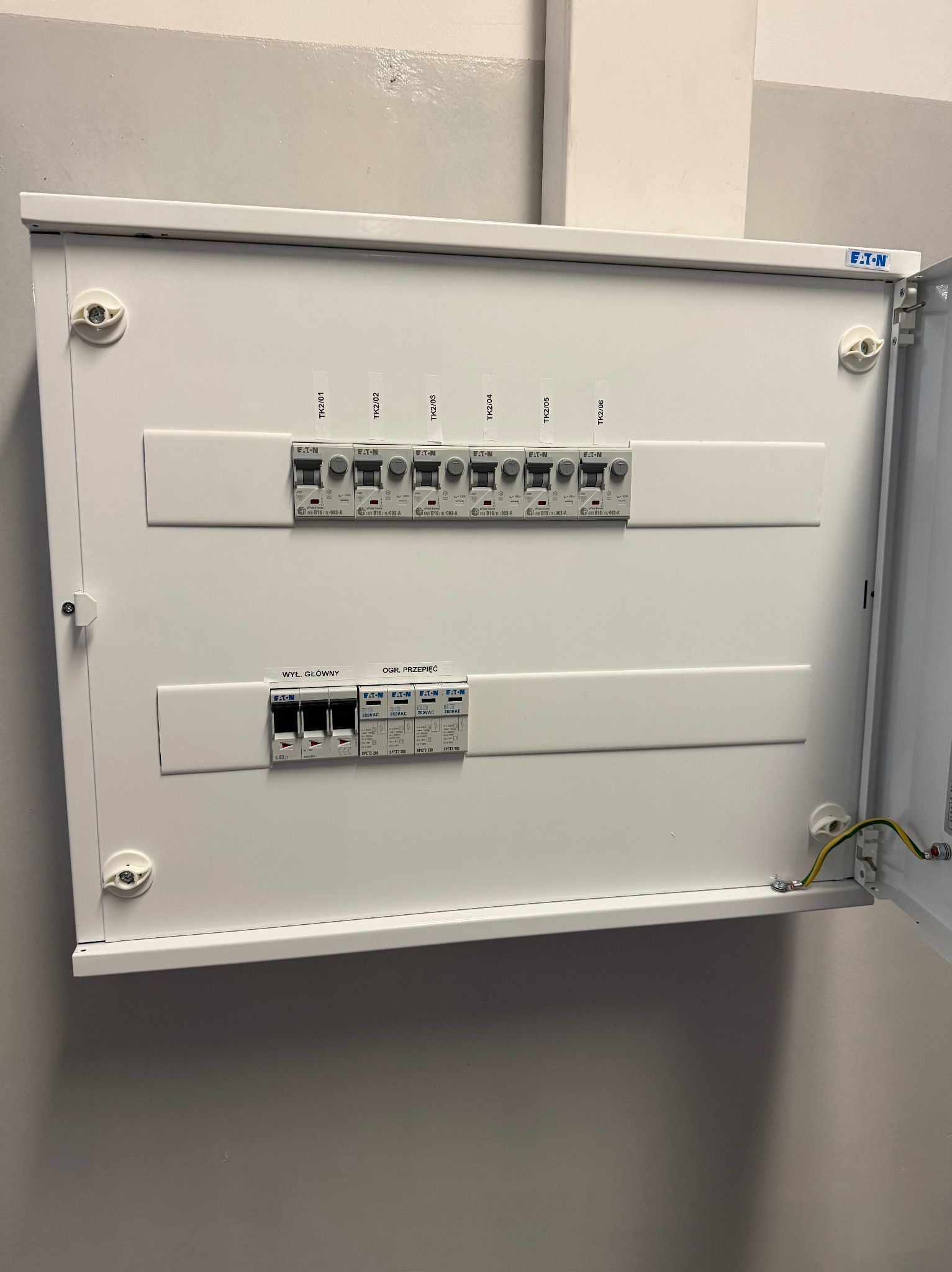 Otwarta, biała skrzynka elektryczna Eaton z bezpiecznikami i ogranicznikiem przepięć, zamontowana na szarej ścianie, widoczne przewody uziemiające.