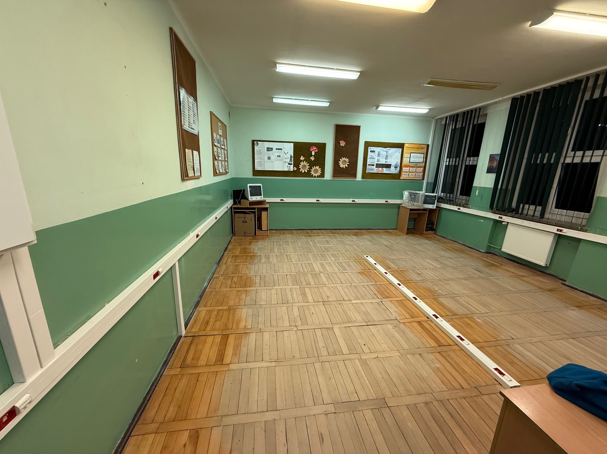 Wnętrze sali gimnastycznej z drewnianą podłogą parkietową, zielonymi ścianami z białym pasem, grzejnikami pod oknami i tablicami informacyjnymi na ścianach.