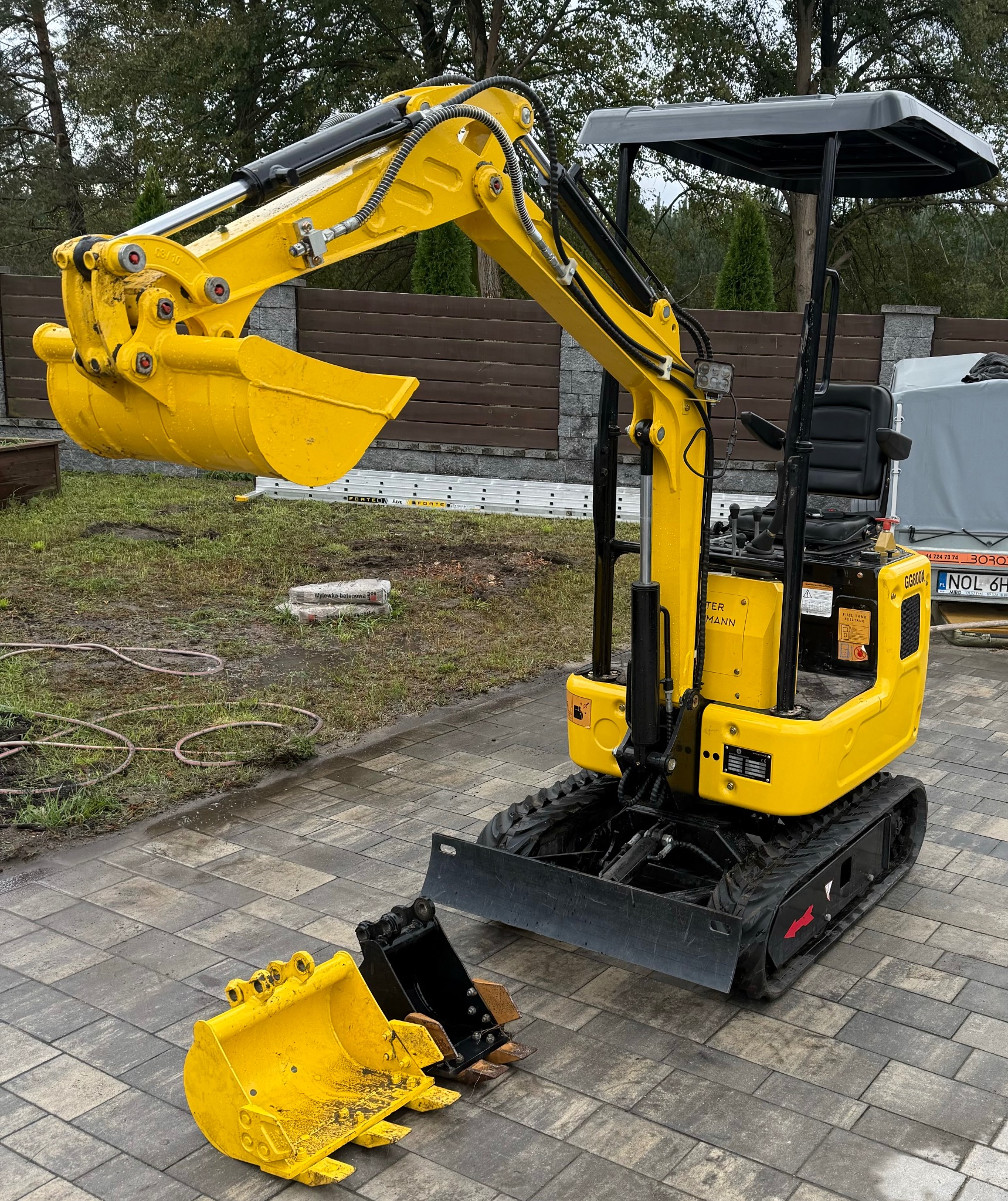 Żółta mini-koparka na brukowanym podjeździe, z dwoma łyżkami obok, widoczne worki z wylewką betonową w tle.