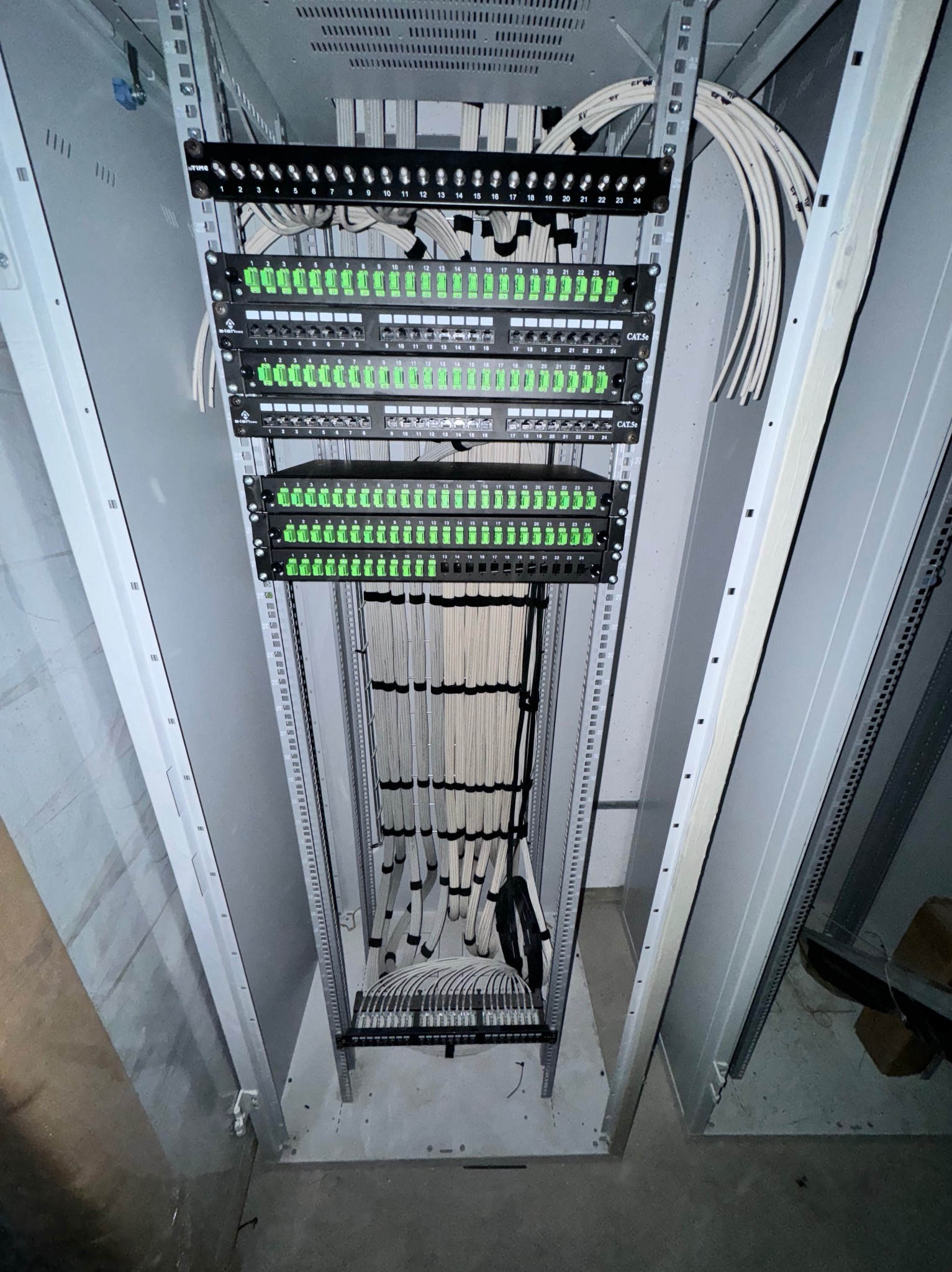 Szafa serwerowa z uporządkowanymi kablami sieciowymi, patch panele z zielonymi portami, widok z dołu w trakcie instalacji.