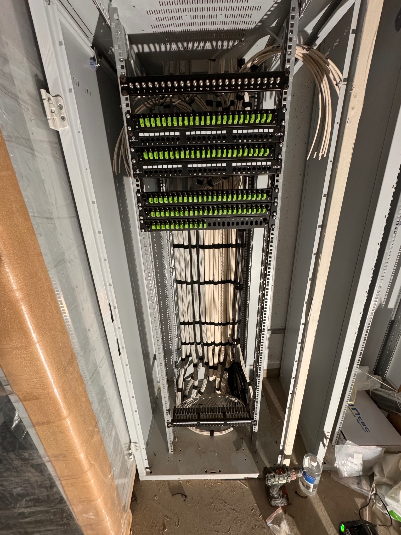 Wnętrze szafy serwerowej w trakcie instalacji okablowania, widoczne panele krosownicze z podłączonymi kablami, narzędzia leżące na podłodze obok szafy.