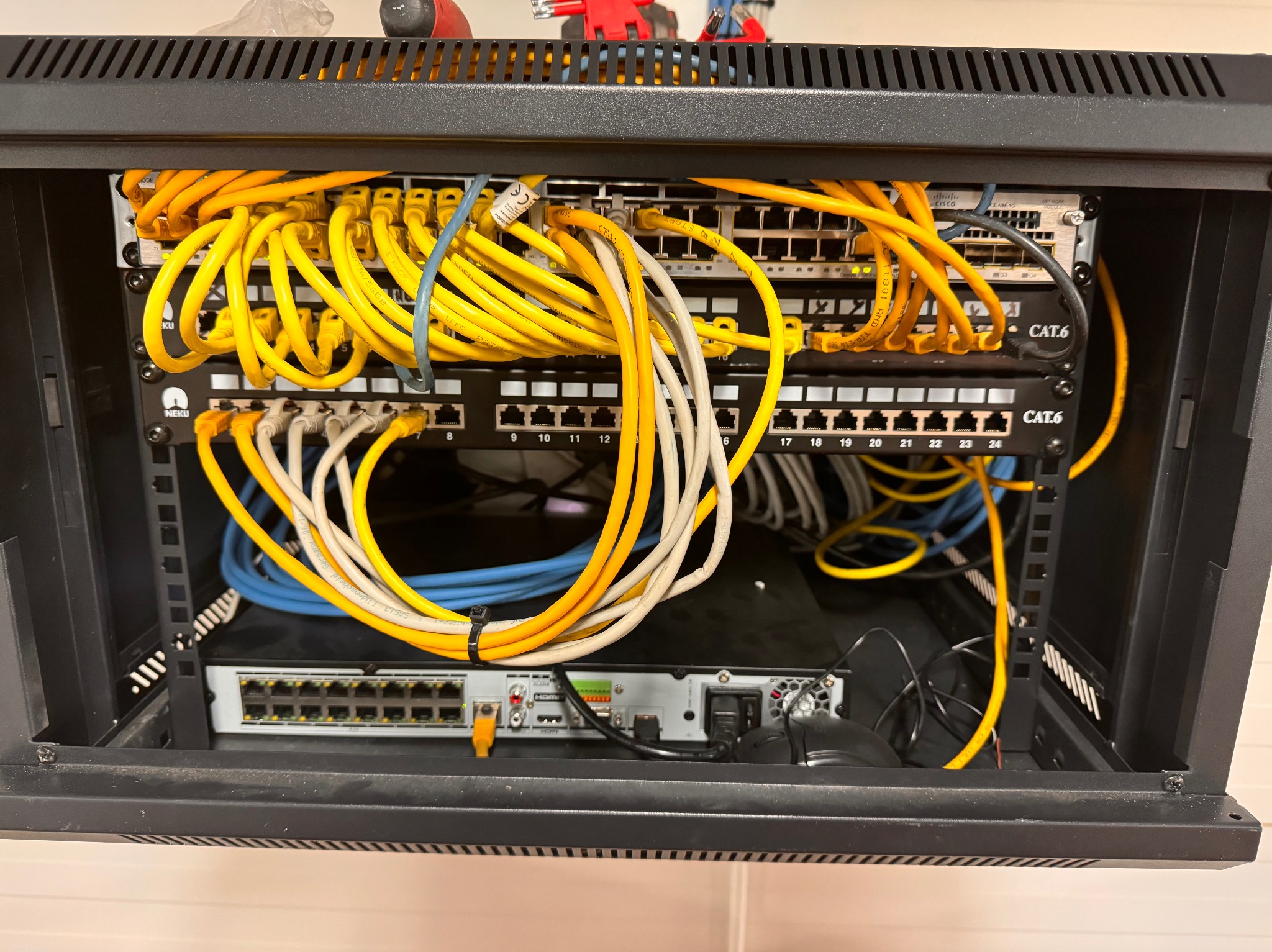 Szafa rack z przełącznikami sieciowymi, patch panelem i okablowaniem CAT6 w kolorach żółtym, białym i niebieskim, widoczne urządzenia Cisco i Neku.