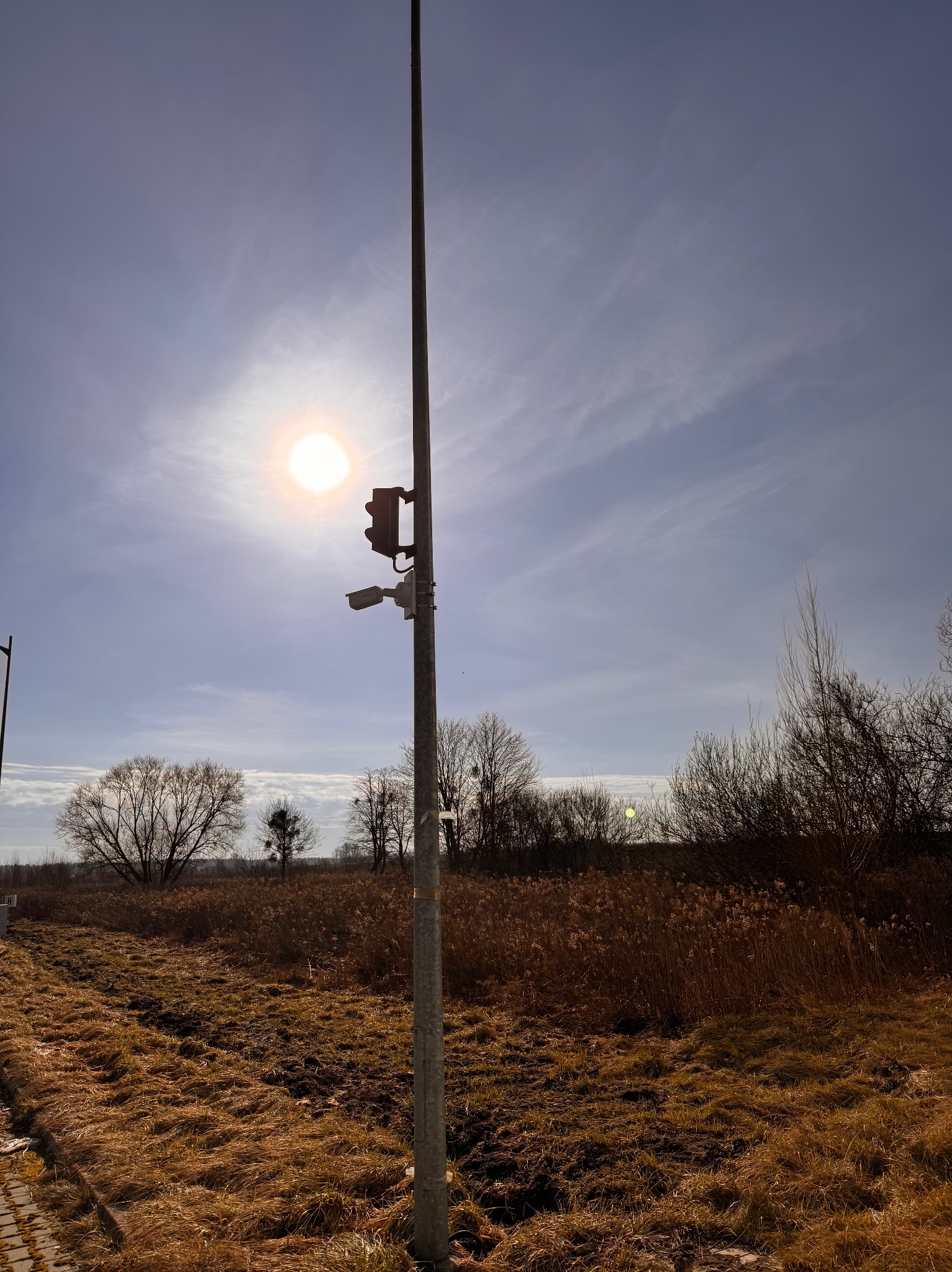Słup z zamontowaną kamerą monitoringu i czujnikiem ruchu na tle jesiennego krajobrazu i jasnego słońca.
