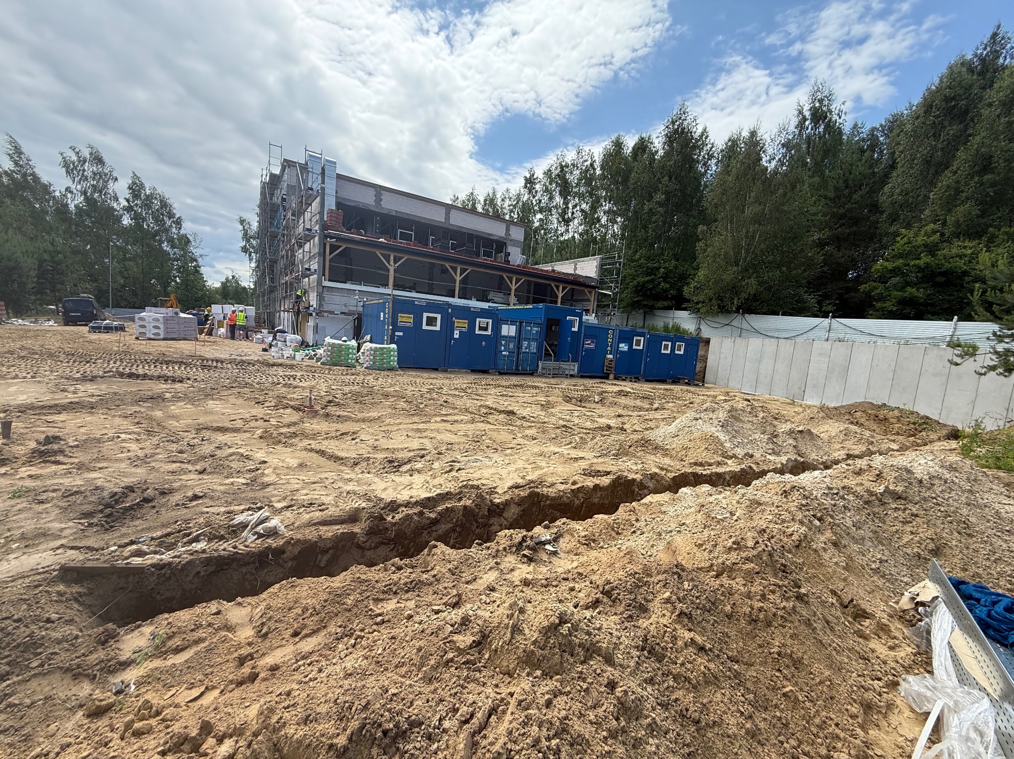 Wykopany rów w ziemi na placu budowy, w tle budynek w trakcie wznoszenia, kontenery budowlane i pracownicy w odblaskowych kamizelkach. Pochmurne niebo.