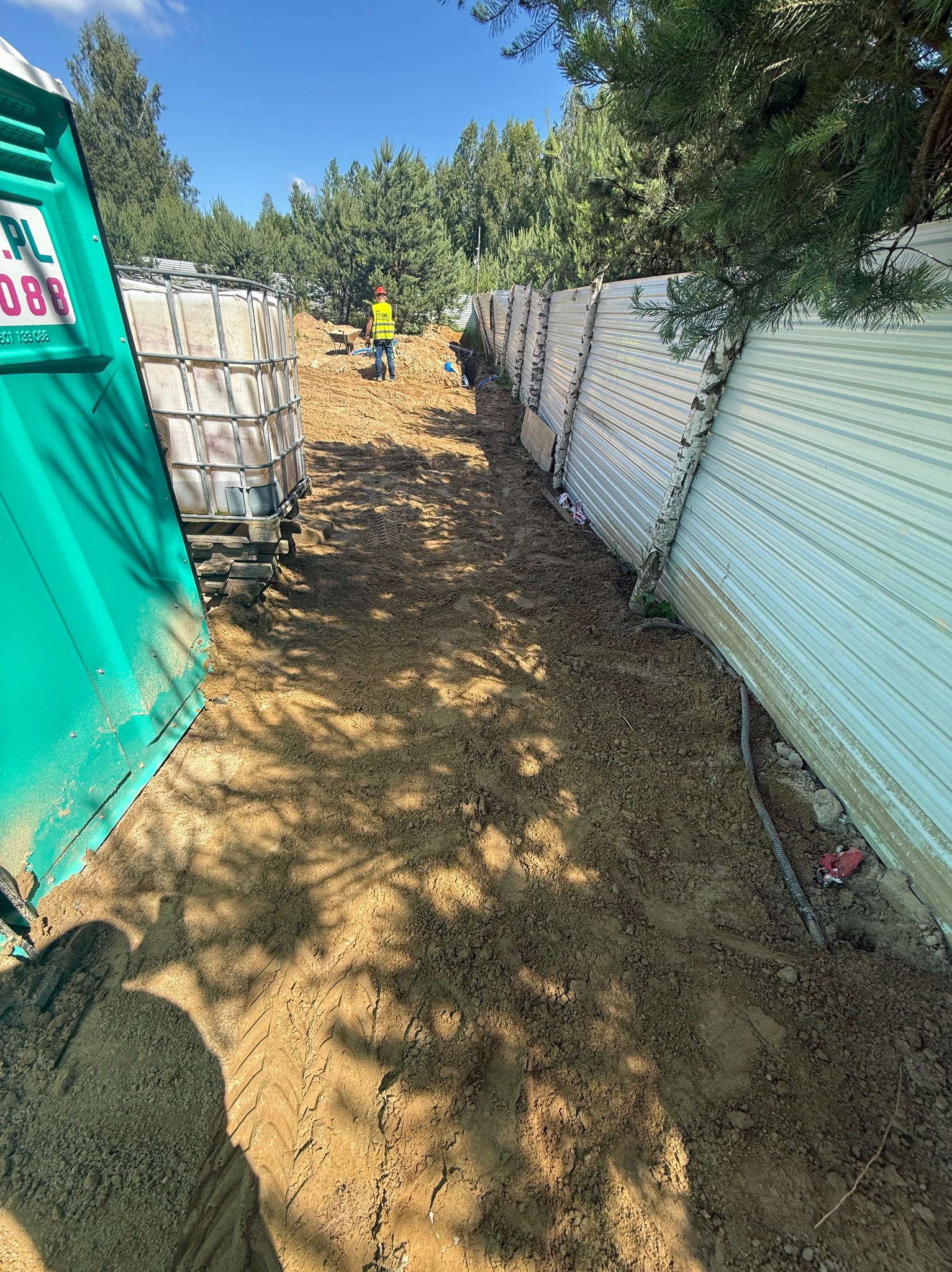 Wykop pod instalacje wzdłuż tymczasowego ogrodzenia z blachy falistej. Widoczny pracownik w odblaskowej kamizelce i fragmenty rur. Ślady opon na ziemi.