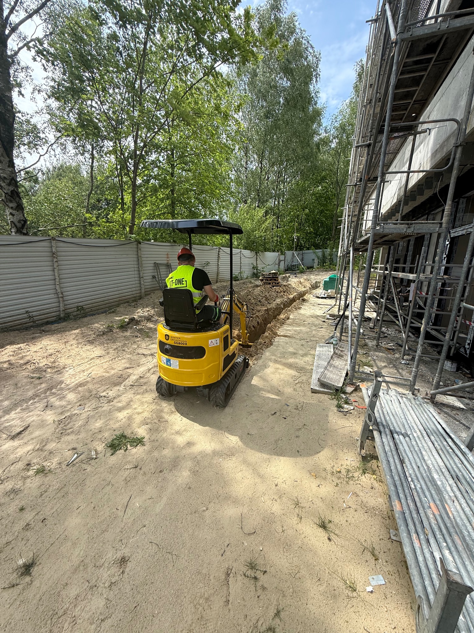 Żółta mini koparka wykonuje wykop w piaszczystej ziemi obok budynku w rusztowaniach, widoczny operator w kamizelce odblaskowej i czerwonym kasku.