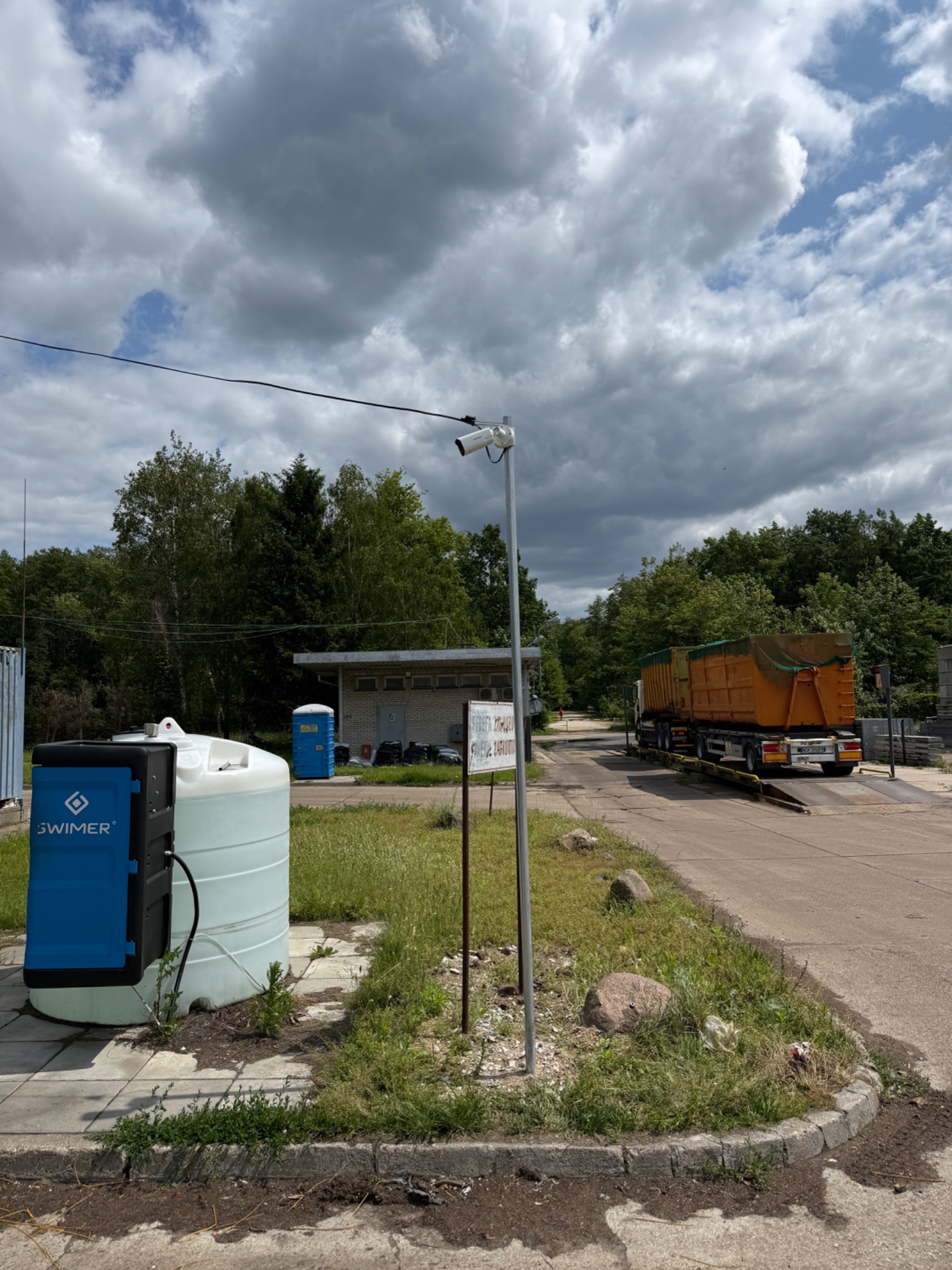 Kamera monitoringu na zewnątrz, nadzorująca wjazd dla ciężarówek; zbiorniki na płyny i budynek w tle, w dzień. System zabezpieczeń i kontroli dostępu na terenie firmy.