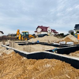 JSB - budujemy Wasz wymarzony dom - Żółta koparka JCB na placu budowy fundamentów domu jednorodzinnego, widoczne zbrojenia i izolacja. W tle zabudowa jednorodzinna i pochmurne niebo.