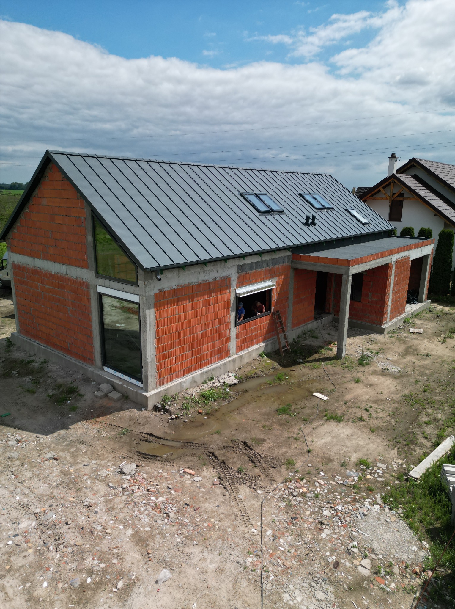 Surowy dom z czerwonej cegły i szarą blachą na dachu, okna wstawione, teren wokół w trakcie przygotowania, widok z góry.