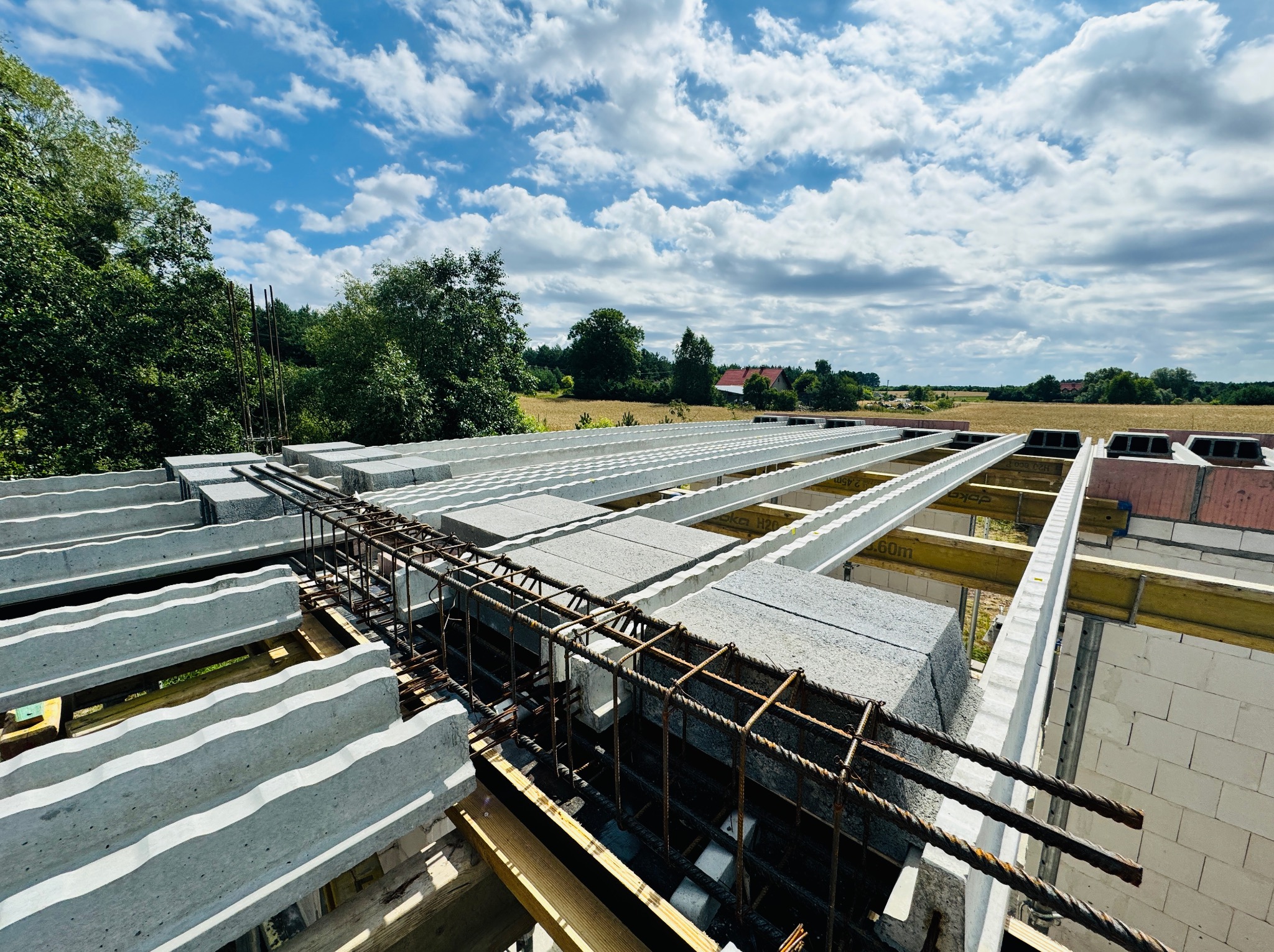 Wieniec stropowy zbrojony stalą na budowie domu, widok z góry, błękitne niebo w tle. Elementy szalunku widoczne. Prace budowlane w toku.