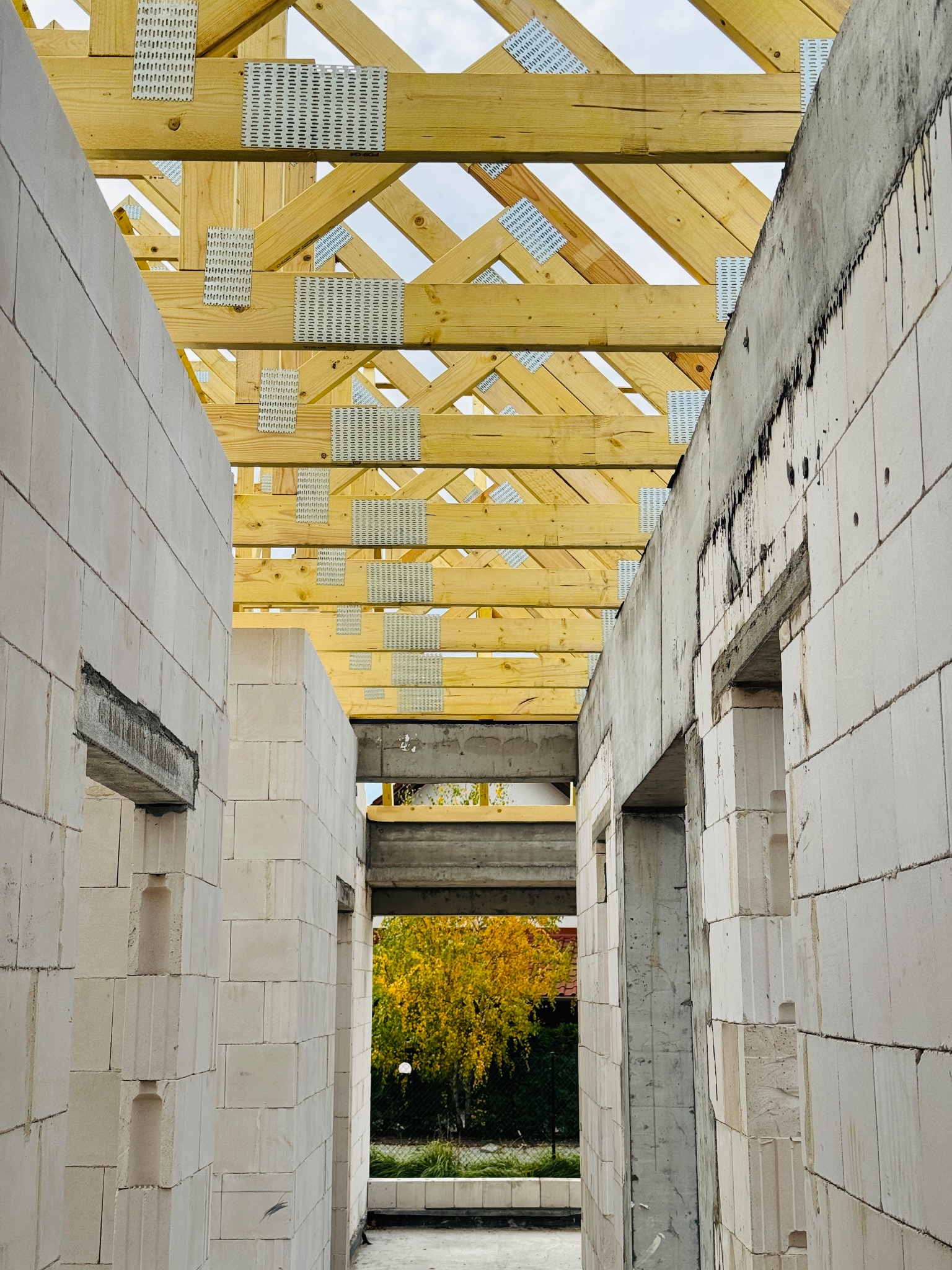Wnętrze domu w trakcie budowy: ściany z białych bloczków, drewniany szkielet dachu z metalowymi łącznikami. W tle widoczne drzewa z żółtymi liśćmi.