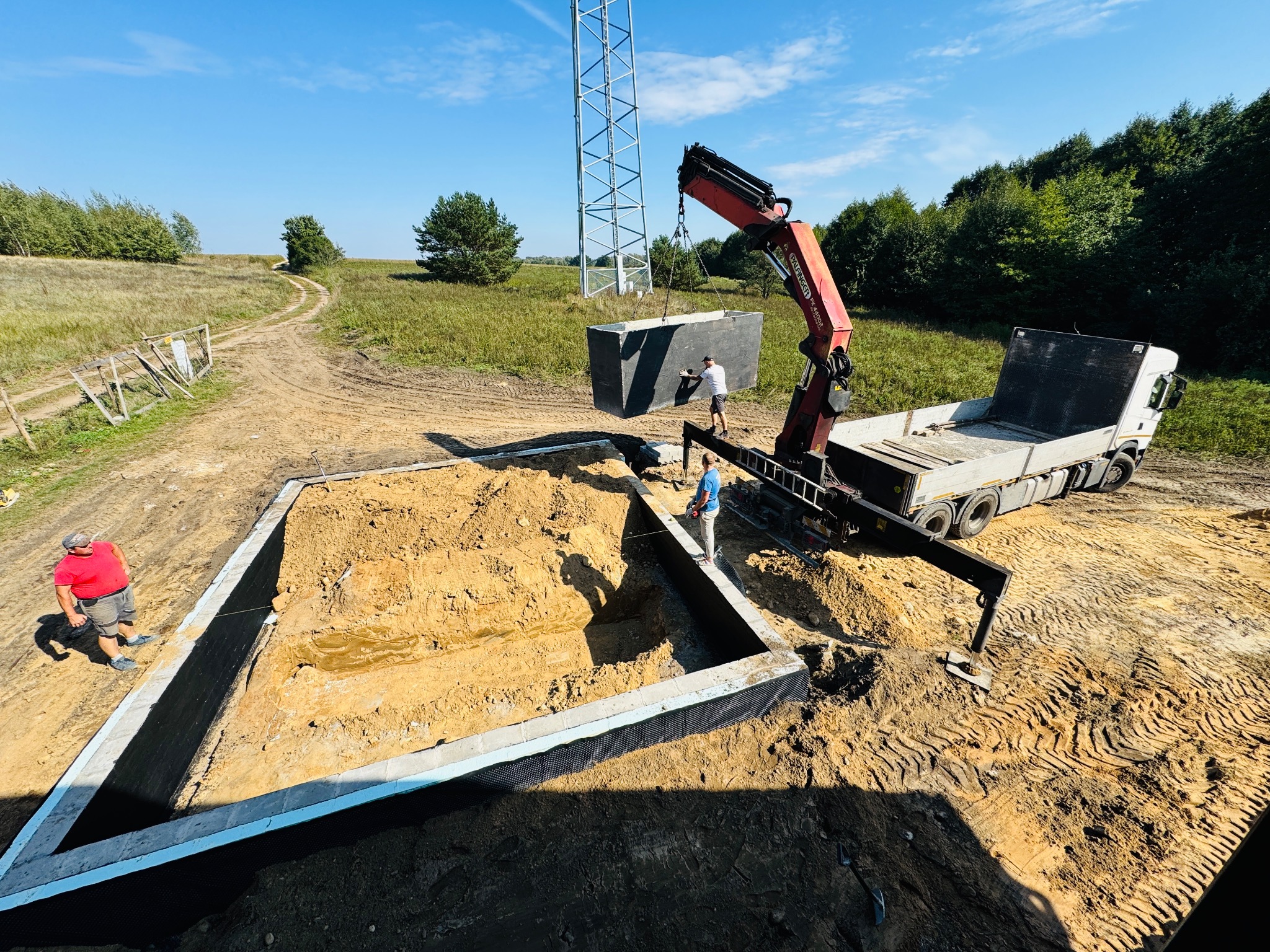 Montaż prefabrykowanego elementu fundamentu za pomocą dźwigu na placu budowy domu jednorodzinnego, w otoczeniu zieleni i wieży telekomunikacyjnej.