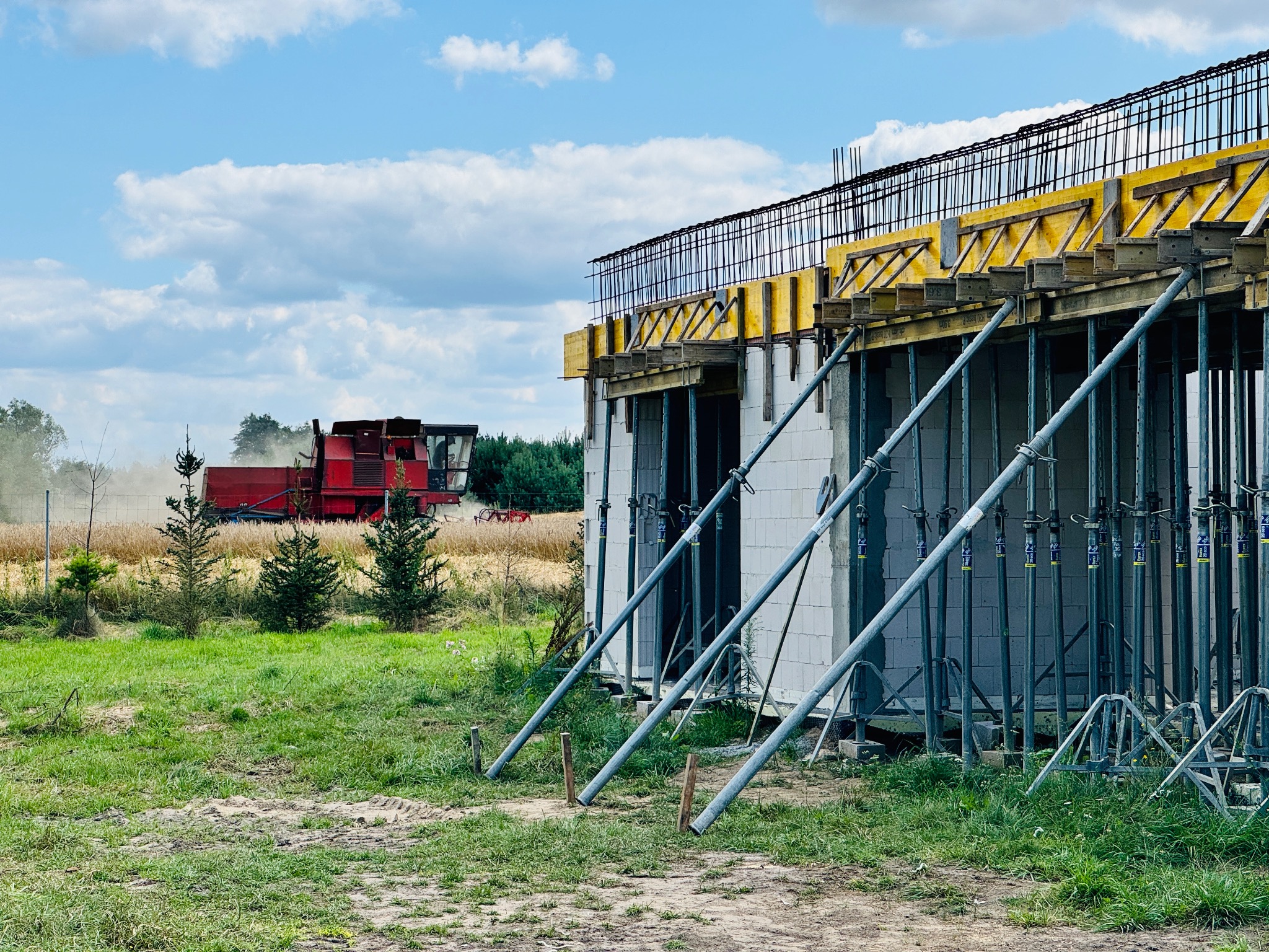 Budowa domu z betonu komórkowego, zbrojenie stropu i szalunki. W tle kombajn zbożowy na polu, błękitne niebo z chmurami.