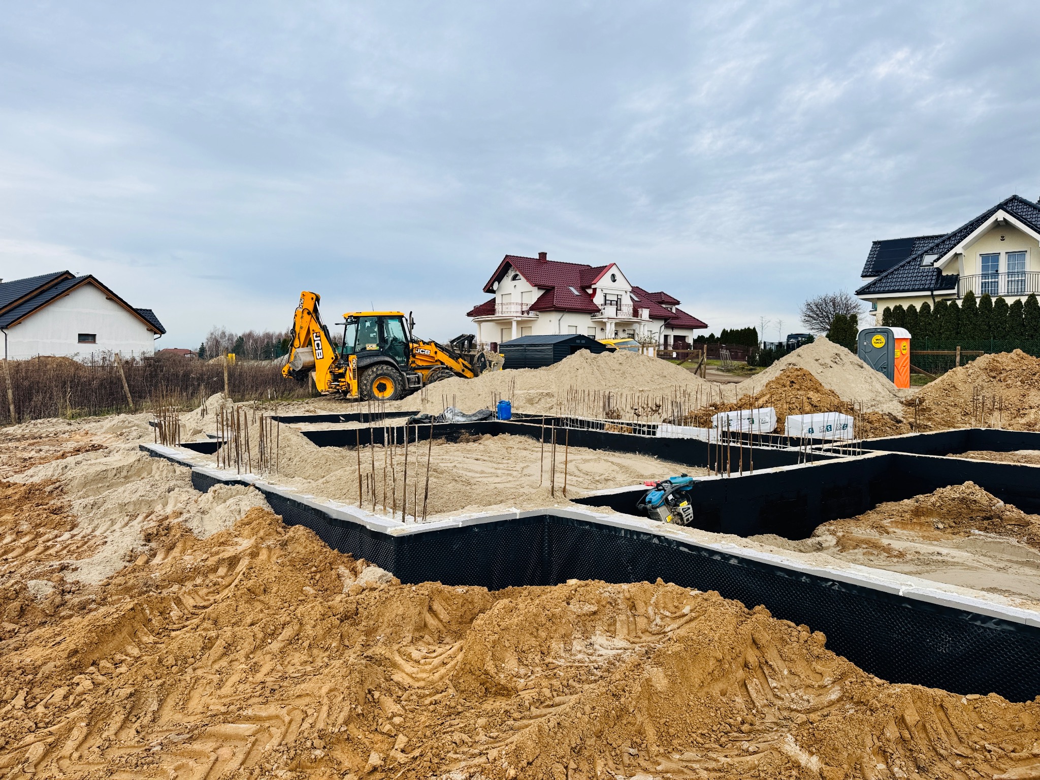 Żółta koparka JCB na placu budowy fundamentów domu jednorodzinnego, widoczne zbrojenia i izolacja. W tle zabudowa jednorodzinna i pochmurne niebo.