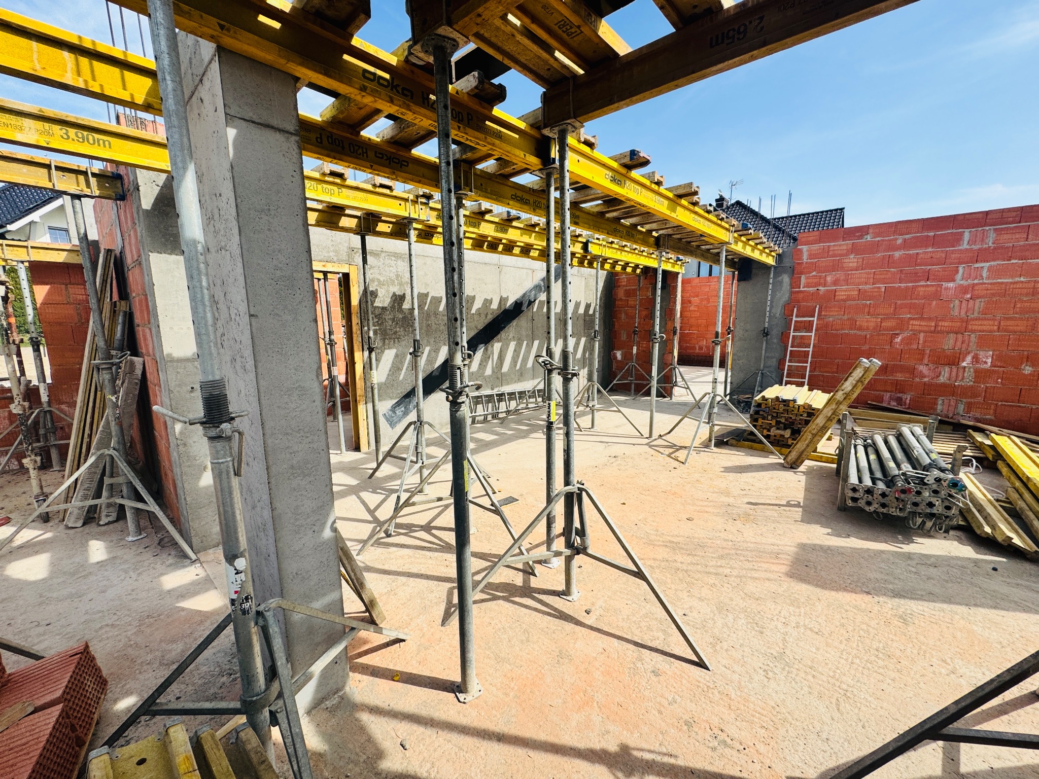Szare betonowe filary i czerwona ściana z cegieł na placu budowy domu, z żółtymi elementami szalunkowymi i podporami. Widoczne niebo i fragment dachu.