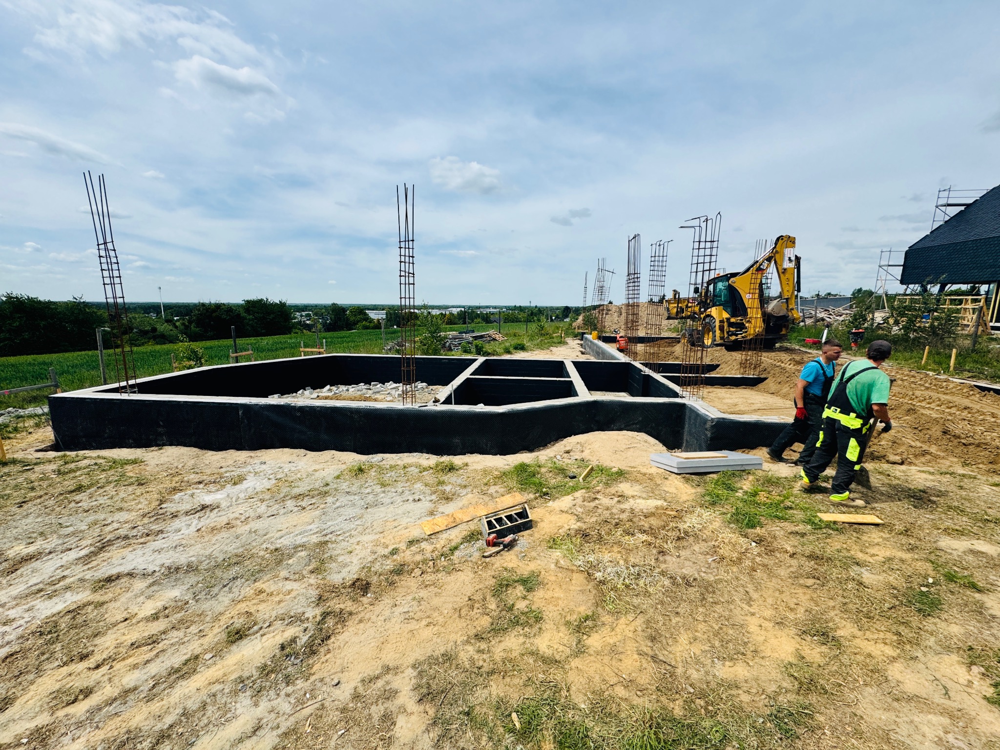 Zalane fundamenty pod budowę domu, zbrojenie pionowe i żółta koparka w tle. Dwóch robotników w strojach roboczych. Widok na okolicę i niebo.