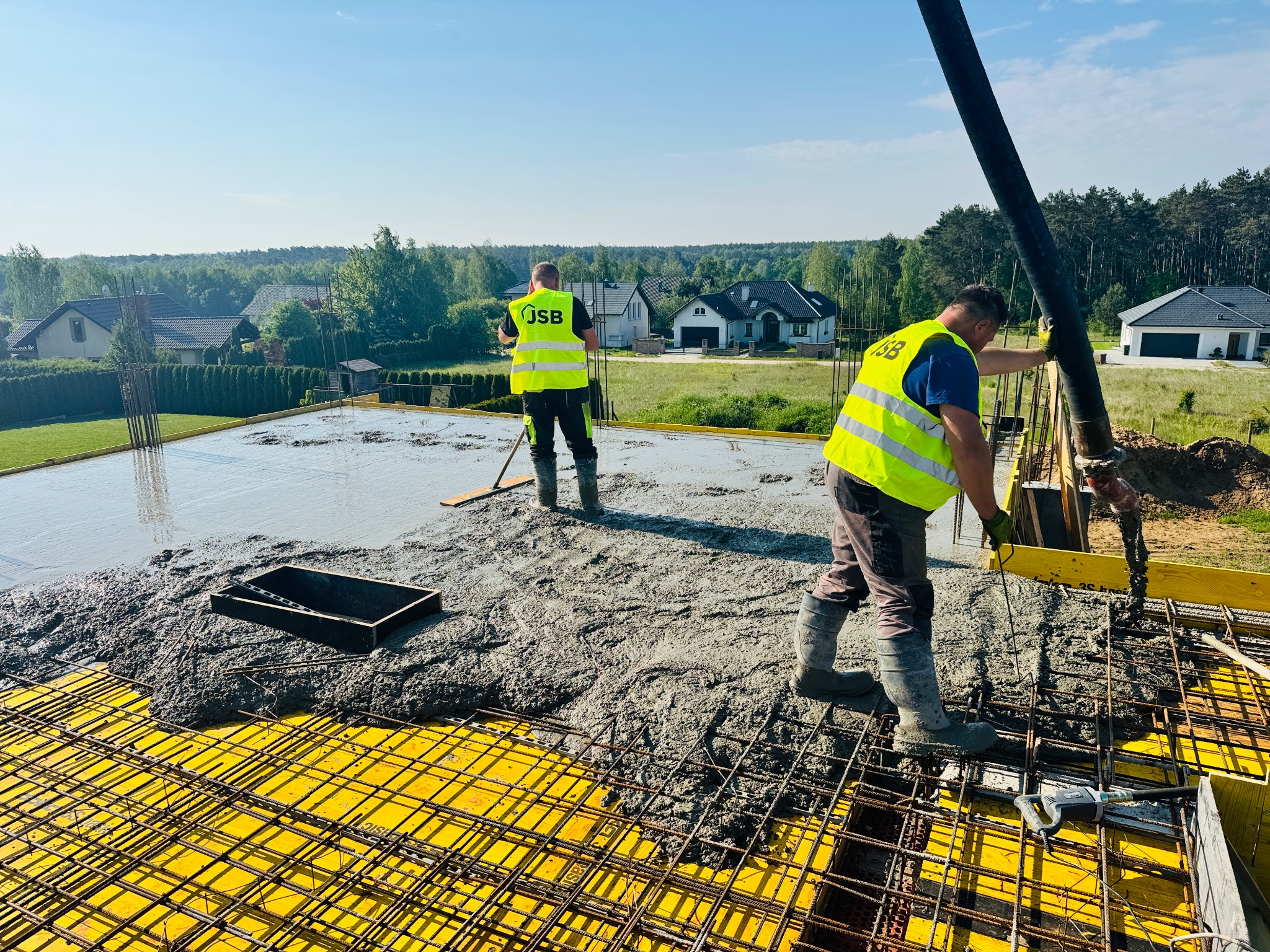 Wylewanie betonu na strop z żółtym szalunkiem i zbrojeniami. Dwóch pracowników w odblaskowych kamizelkach. W tle domy i las.