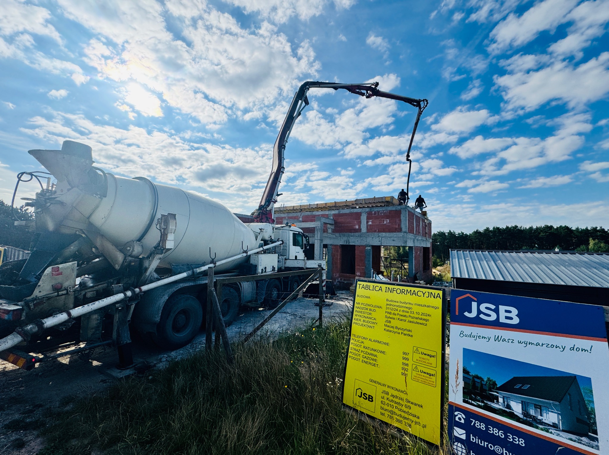 Betonomieszarka pompuje beton na budowę domu z czerwonej cegły. Widoczni robotnicy. Tablica informacyjna i reklama firmy JSB na pierwszym planie.