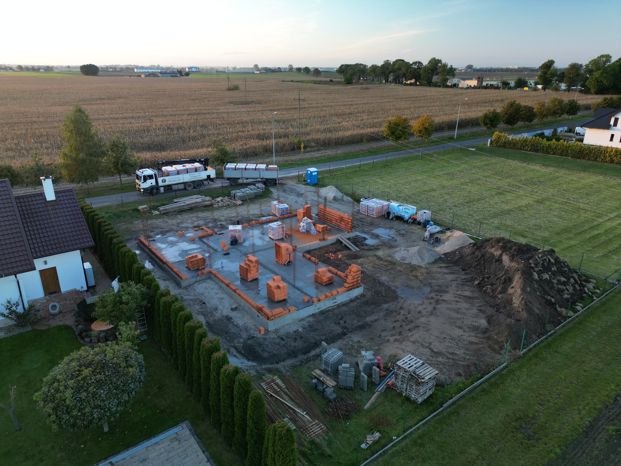 Widok z lotu ptaka na plac budowy domu jednorodzinnego w Libartowie, z widocznymi fundamentami, częściowo wymurowanymi ścianami z czerwonej cegły, dostawą materiałów budowlanych ciężarówką...