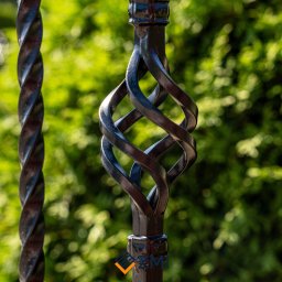 Zakład Metalowo Instalacyjny Dębica - Fragment czarnej, metalowej balustrady z ozdobnym elementem skręconym w kształt koszyczka, na tle zieleni.