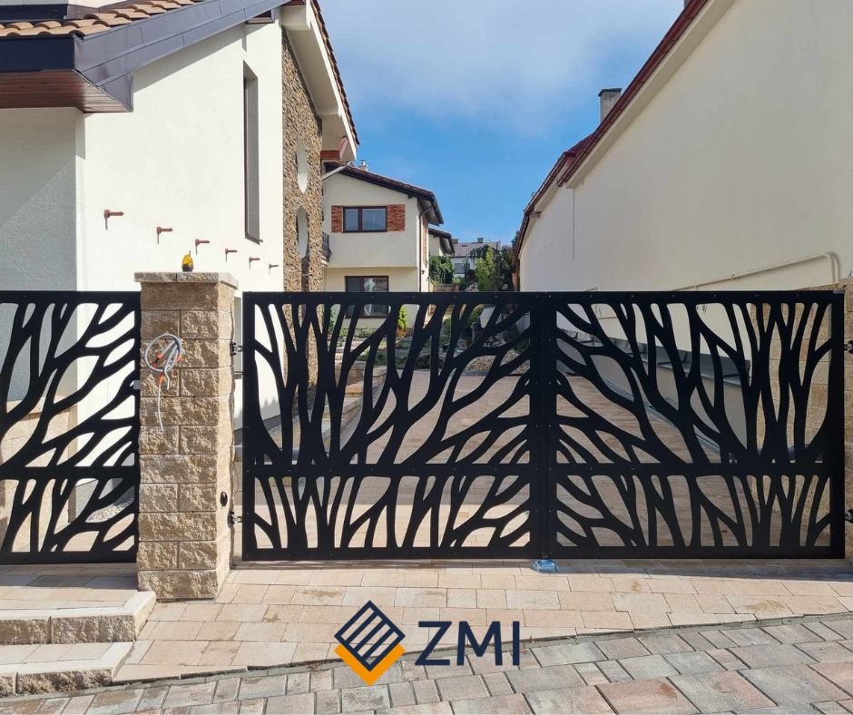 Nowoczesna, czarna brama wjazdowa z laserowo wycinanym wzorem gałęzi na tle domów jednorodzinnych, z kamiennymi słupkami i brukowanym podjazdem.