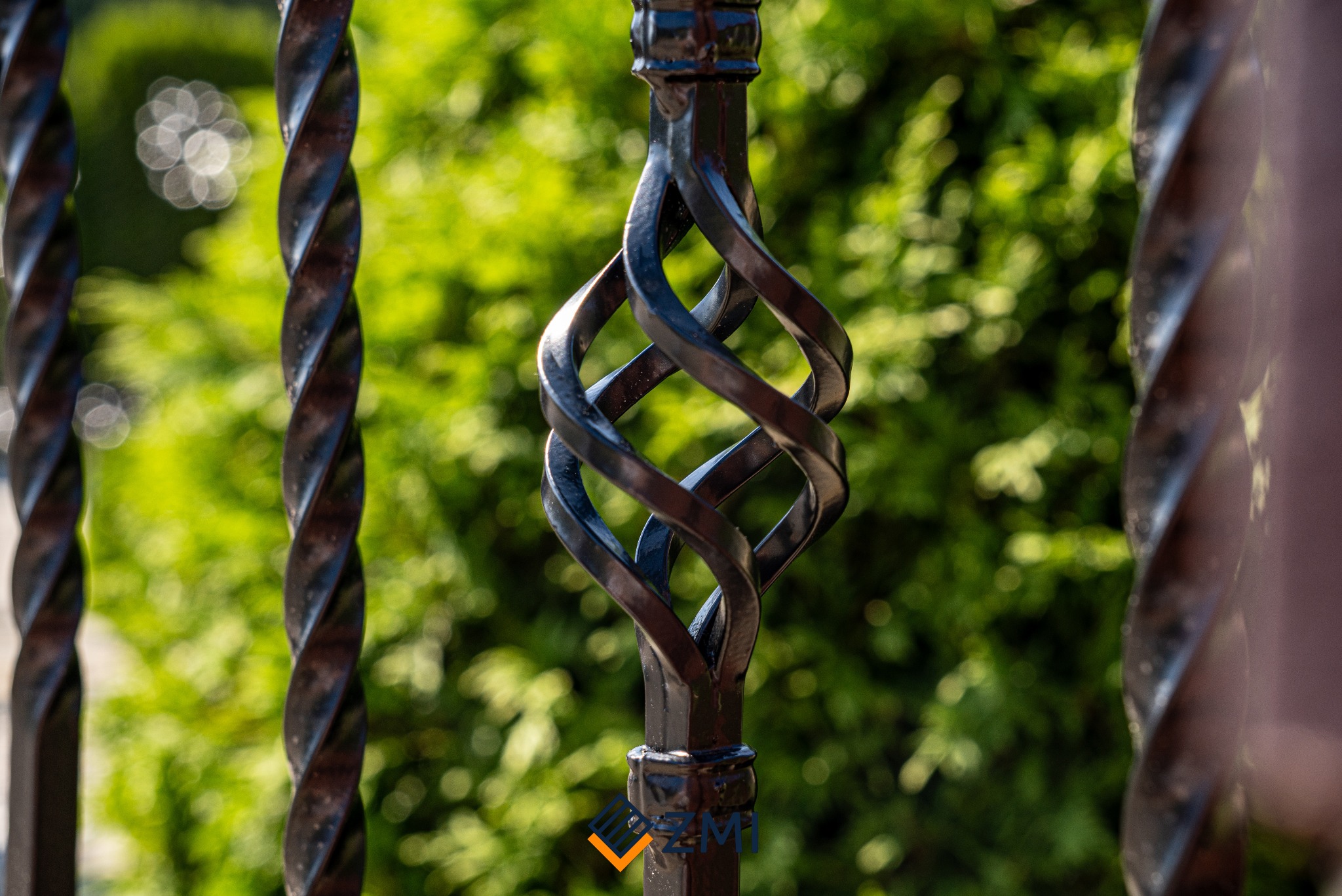 Fragment czarnej, metalowej balustrady z ozdobnym elementem skręconym w kształt koszyczka, na tle zieleni.