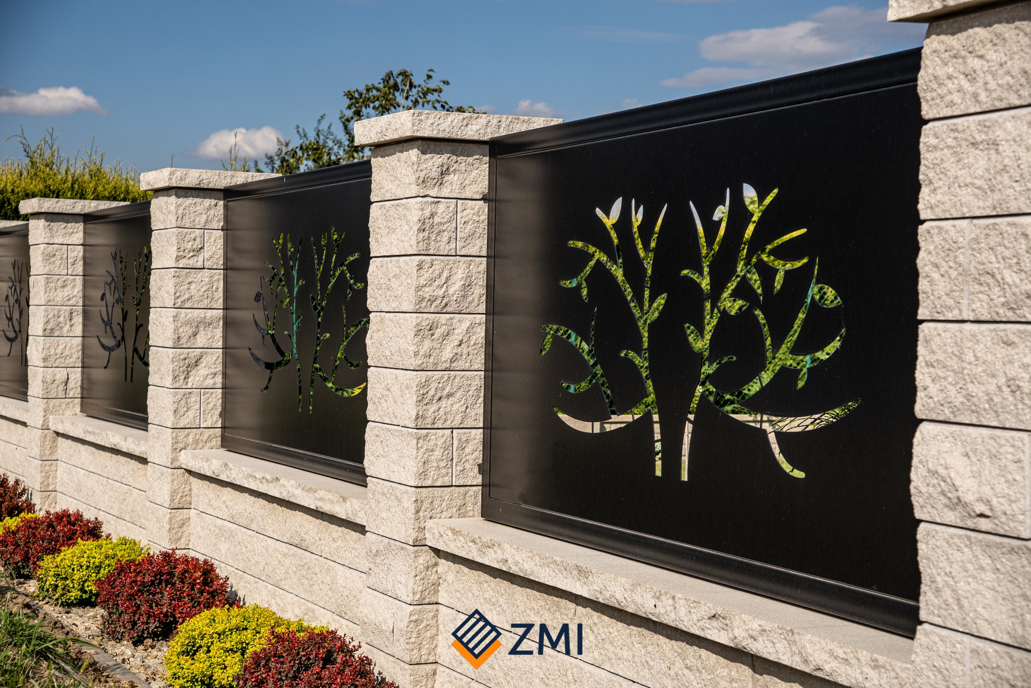 Nowoczesne ogrodzenie z czarnymi panelami z motywem drzewa, osadzone na kamiennych słupkach, z rabatą z kolorowych krzewów u podstawy.