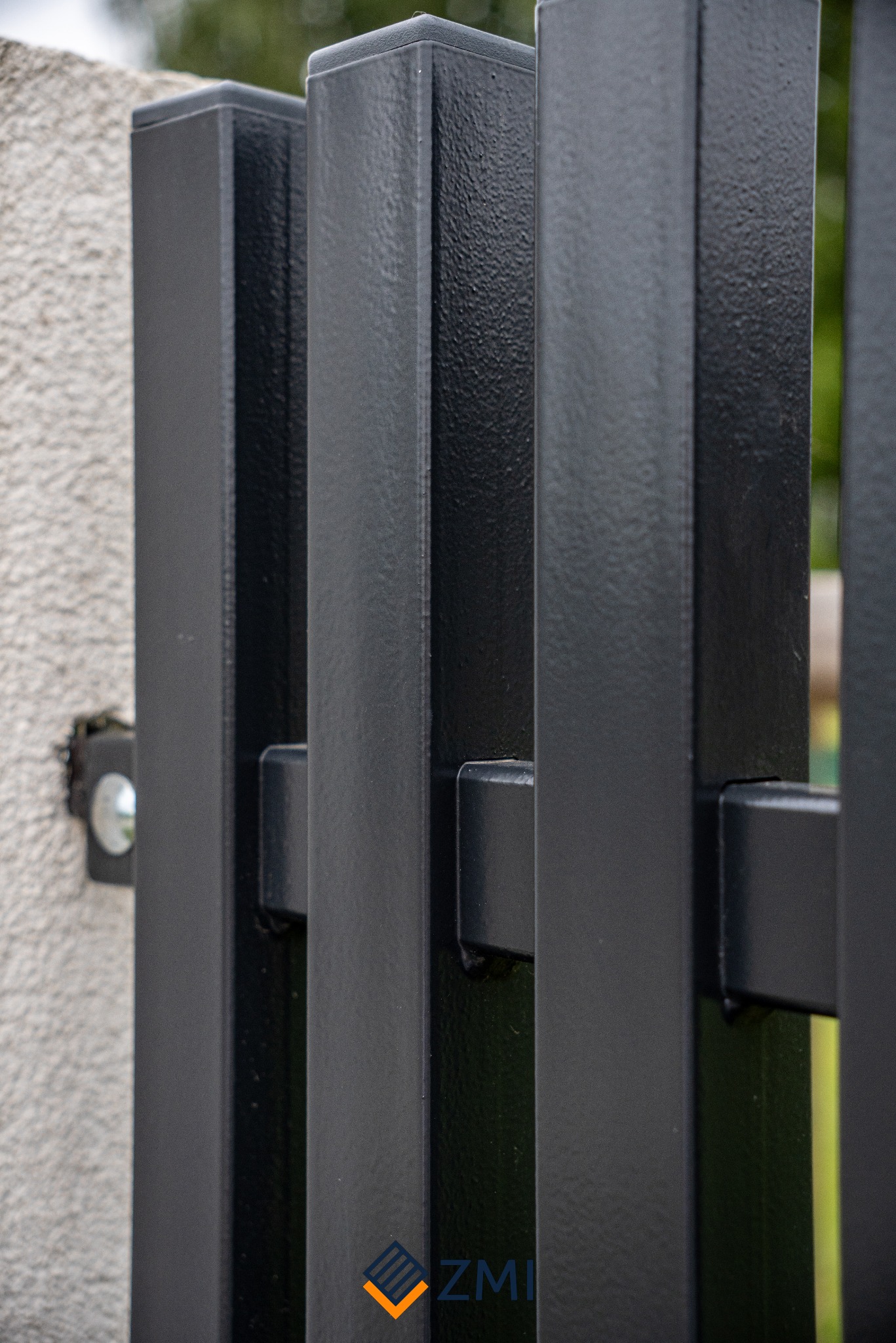 Fragment czarnego, metalowego ogrodzenia z profilowanymi słupkami, zamontowanego przy szarym, teksturowanym murze.