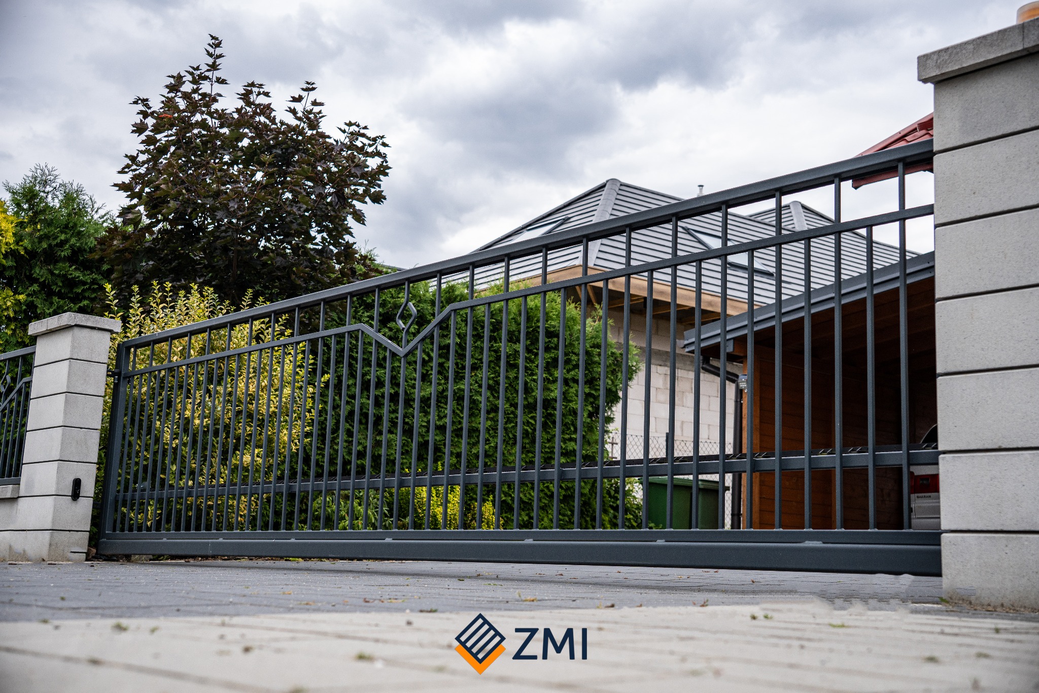 Przesuwna brama wjazdowa z metalowymi sztachetami, widok z poziomu gruntu na tle domu i zieleni.