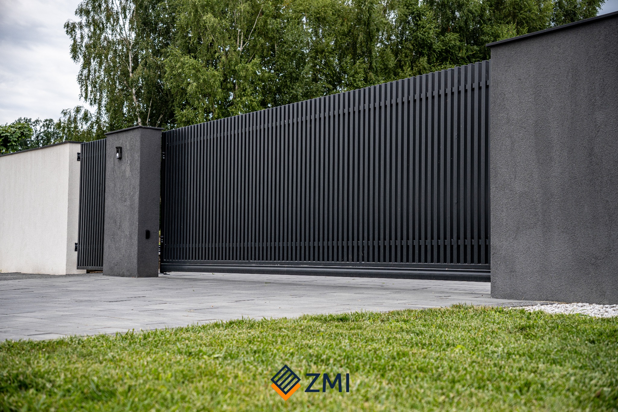 Nowoczesna, przesuwna brama w kolorze antracytowym z pionowymi panelami, osadzona między dwoma masywnymi, szarymi filarami, widoczna z poziomu trawnika, z logo ZMI w prawym dolnym rogu.