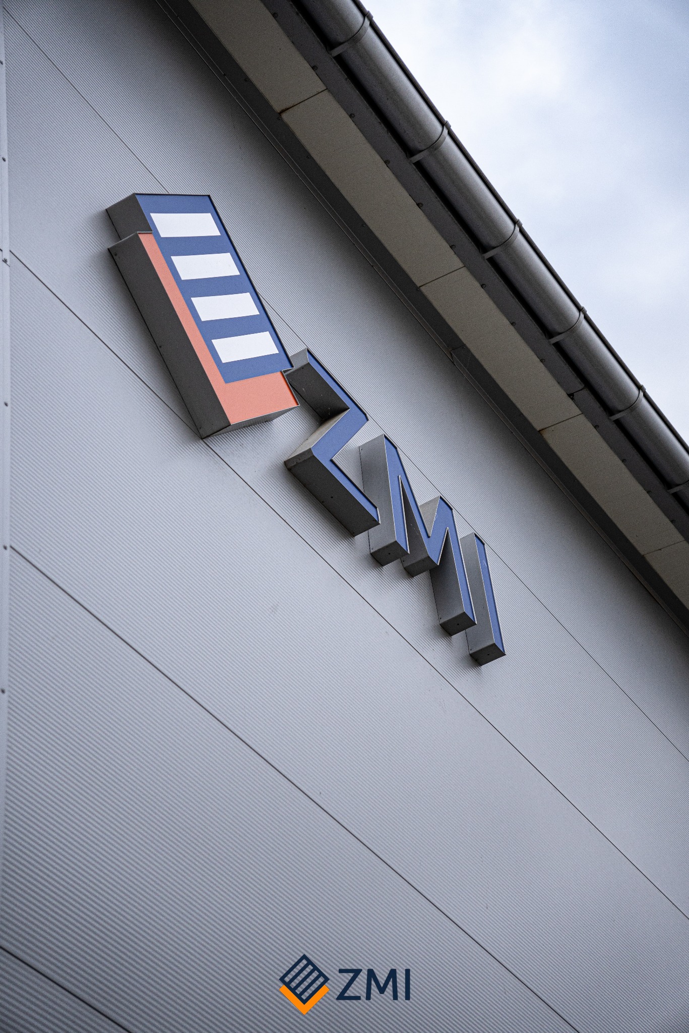 Elewacja budynku z logo firmy ZMI, składającym się z geometrycznego symbolu i liter w kolorach niebieskim, pomarańczowym i białym, widoczne rynny dachowe.