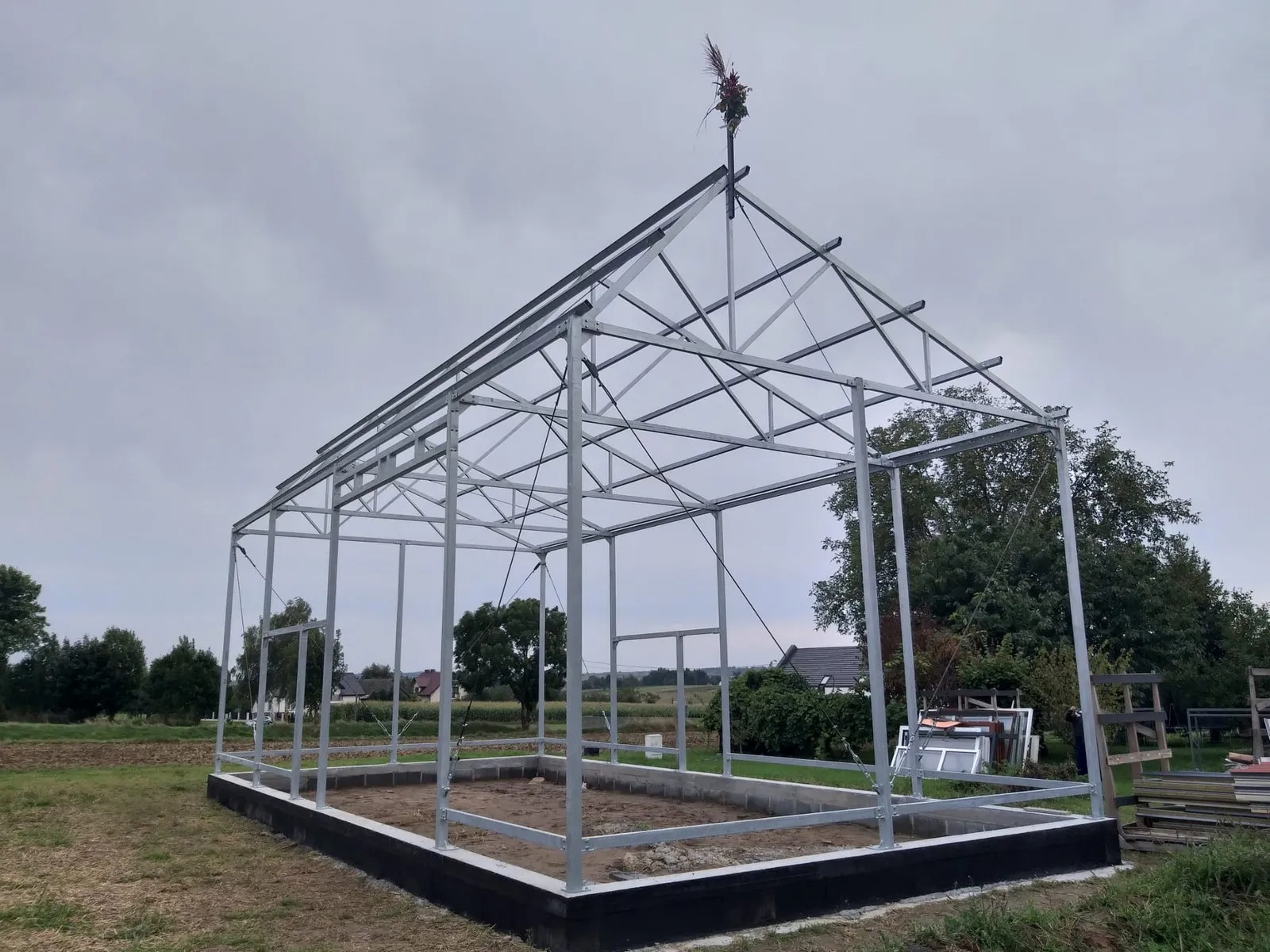 Konstrukcja szkieletowa hali w trakcie budowy, z wieńcem na szczycie, na tle pochmurnego nieba i zieleni.